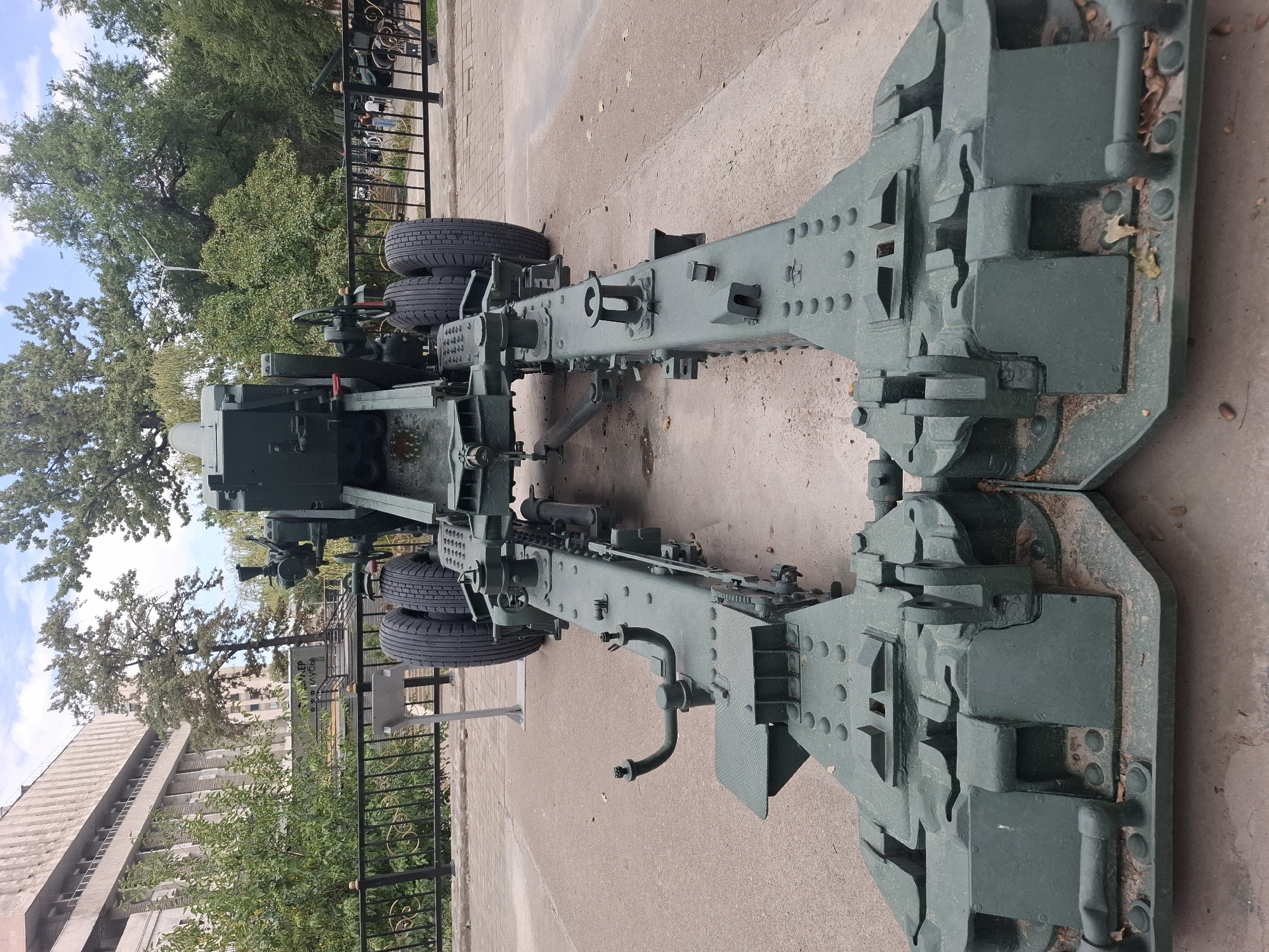 This is a full-shot, eye-level, vertical, outdoor photograph of a large, dark green artillery piece parked on a paved area. The artillery piece is positioned in the foreground, taking up most of the frame. It has two large tires, a long, segmented barrel, and a complex mechanical base with what appear to be outriggers that are spread wide on the ground. The object is a static display.
In the background, there is a black metal fence with ornamental scrollwork. Behind the fence, there are several mature trees with green foliage and a large, modern building with a light-colored facade and multiple levels of windows. The sky is partly cloudy. The ground in front of the artillery piece is paved with light brown square tiles. There are some fallen leaves and acorns scattered on the ground.
The artillery piece is a prominent display item. No people are visible in the immediate vicinity of the artillery piece, but some figures can be seen in the distance behind the fence, suggesting this is a public space. The overall impression is of a monument or historical exhibit.
This artillery piece is located in Almaty, Kazakhstan, specifically at the Museum of the Armed Forces of the Republic of Kazakhstan.