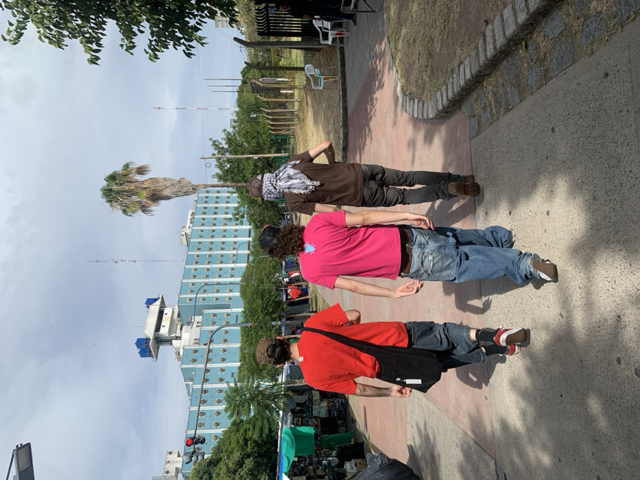 Three men walk along a paved path. The first wears a brown shirt and checkered scarf, the second a pink shirt and jeans, and the third a red shirt with a black bag. A tall building with many windows and a palm tree are in the background. There are trees to the left of the building and a fence with a grassy area to the right.