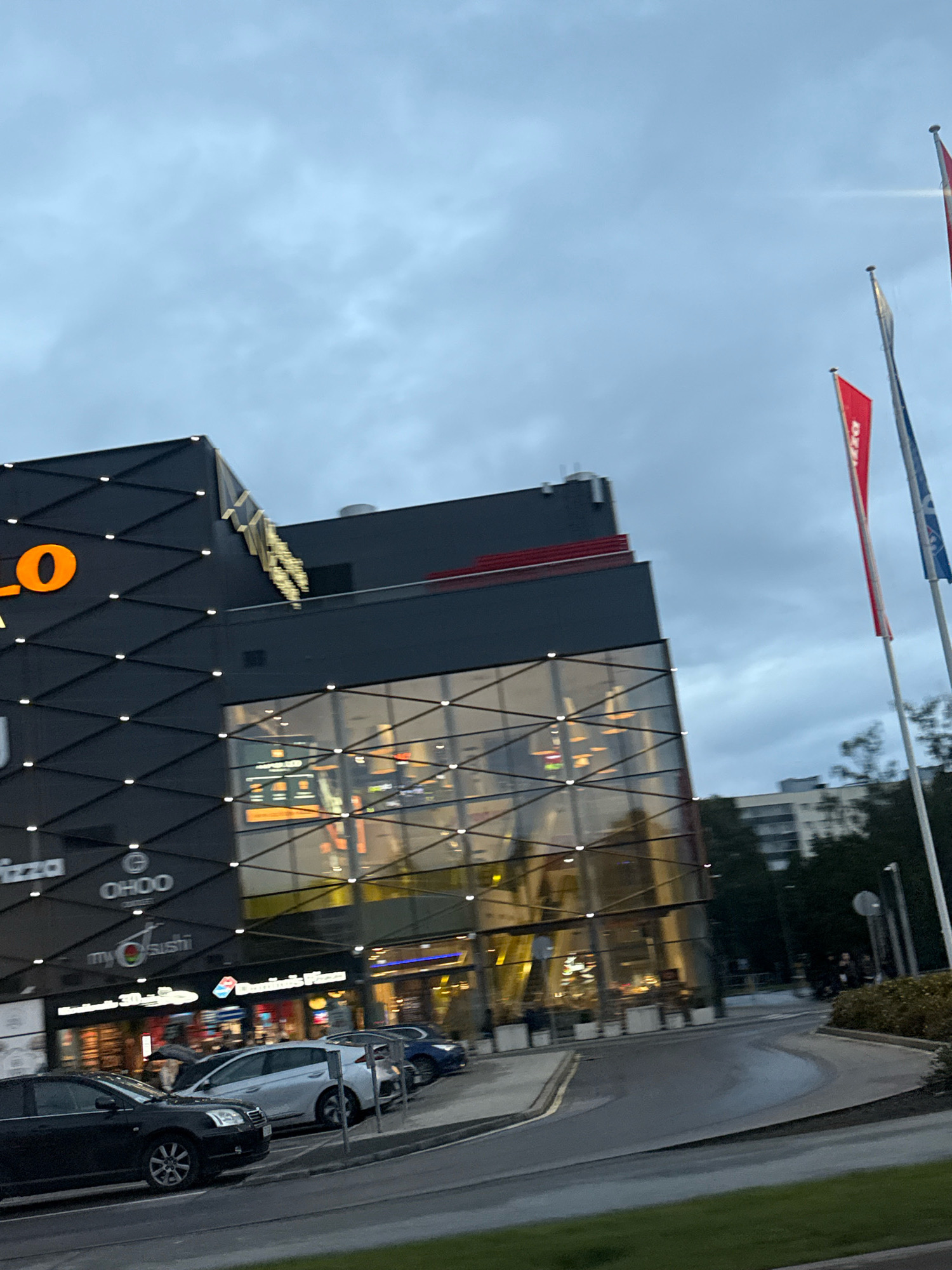 The image shows a modern building complex with a black facade featuring a geometric pattern and illuminated dots. The building has large glass windows reflecting the overcast sky. Signage visible includes "LO" in large orange letters, a golden abstract logo, "pizza," "OHOO," "my sushi," and "Domino's Pizza." Several cars are parked in front of the building, including a black sedan, a silver hatchback, and a dark blue sedan. Three flags are flying on poles to the right of the building; two are red with white text, and one is blue. The scene appears to be in an urban environment, likely a commercial area. The overall impression is of a shopping center or a complex with multiple businesses. Based on the signage, this location is likely the Rocca al Mare shopping center in Tallinn, Estonia.