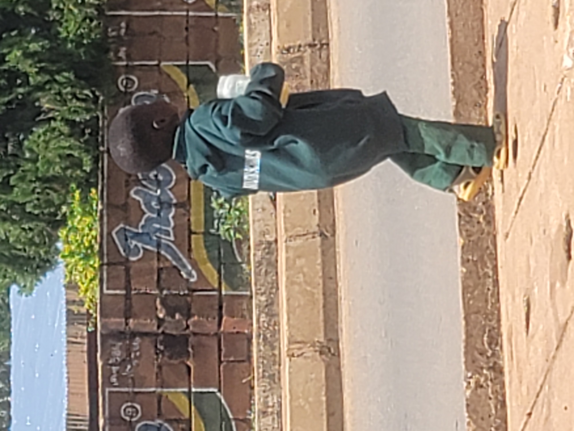 In Barakin Lerawi, Nigeria, a young child walks along a paved sidewalk next to a road. The child is wearing a dark green hooded sweatshirt and matching green pants, with yellow sandals. The sweatshirt has white lettering on the back that reads "MARSKILLS." The child is carrying a white plastic cup or bottle in their right hand. Behind the child is a brick wall decorated with graffiti. The wall is made of brown bricks and features stylized lettering in white and yellow, as well as what appears to be a drawing of a banana. Green foliage is visible above the wall. The road to the left of the sidewalk is grey asphalt. A concrete barrier separates the sidewalk from the road. The lighting suggests it is daytime.