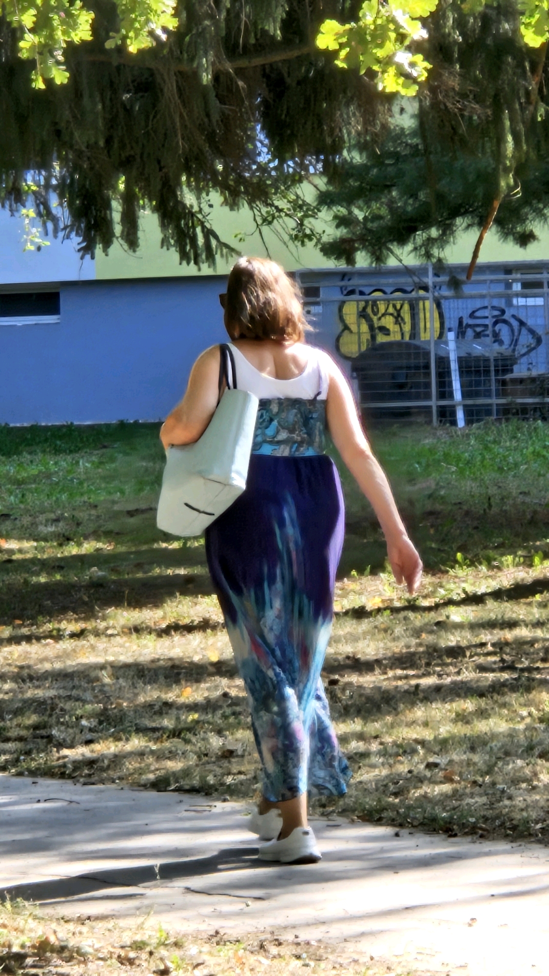 A woman walks away from the camera, her back to the viewer, on a paved path in a grassy area. She has shoulder-length brown hair and wears a white tank top with a blue and purple tie-dye maxi skirt. She carries a light blue tote bag over her left shoulder. The background shows a blue and yellow building with graffiti on a fence. The scene is illuminated by sunlight, casting shadows on the path. The woman is walking in the direction of the building. The location is identified as Prešov, Slovakia.
