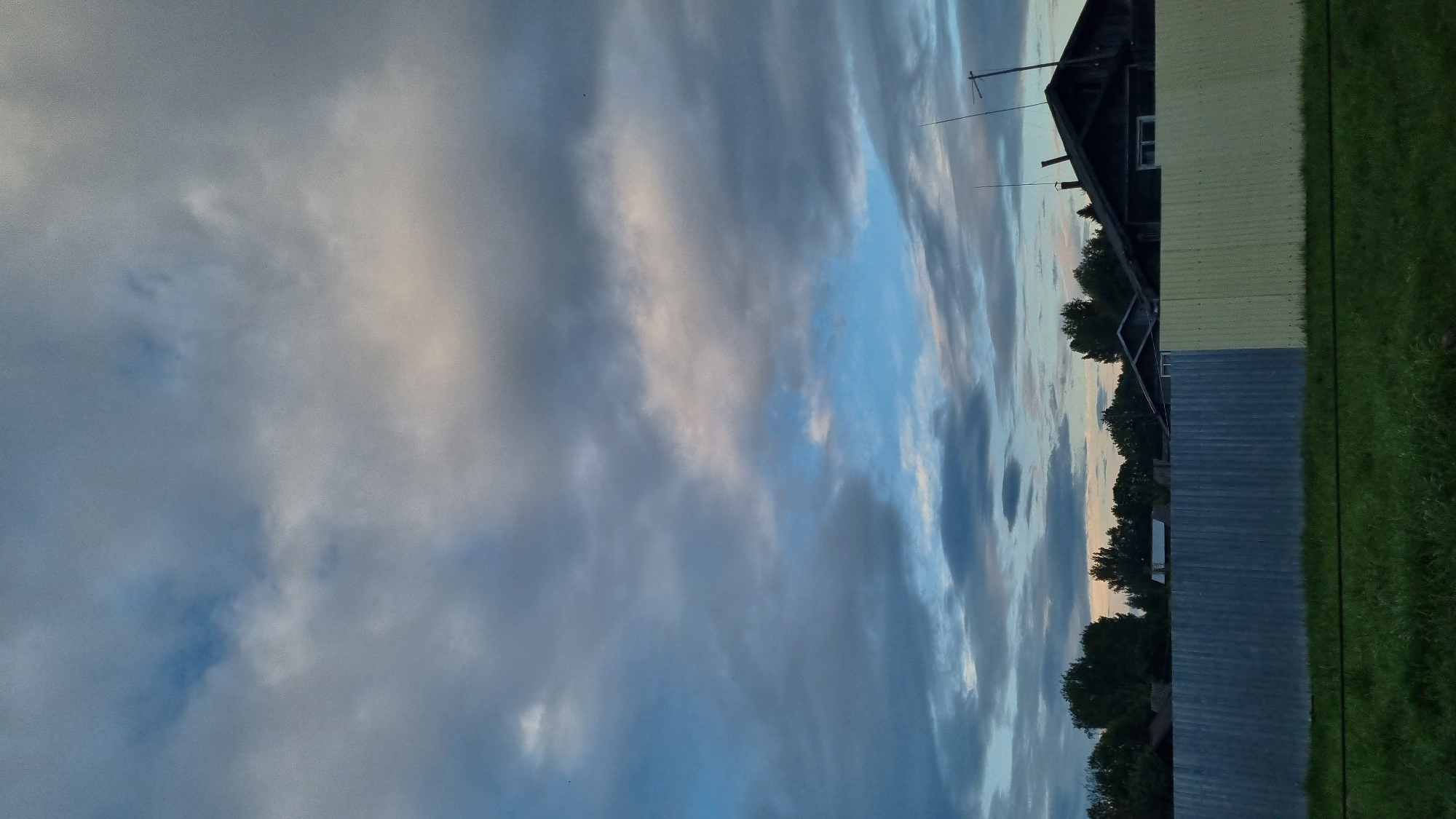 The image shows a fence in the foreground, with a portion of it colored gray and the other yellow. Behind the fence, there are trees, houses, and a cloudy sky with patches of blue. There is also a patch of green grass in front of the fence. The image is taken from an outdoor setting. The scene appears to be a residential area. The location is identified as Vysokoe, Russia.
