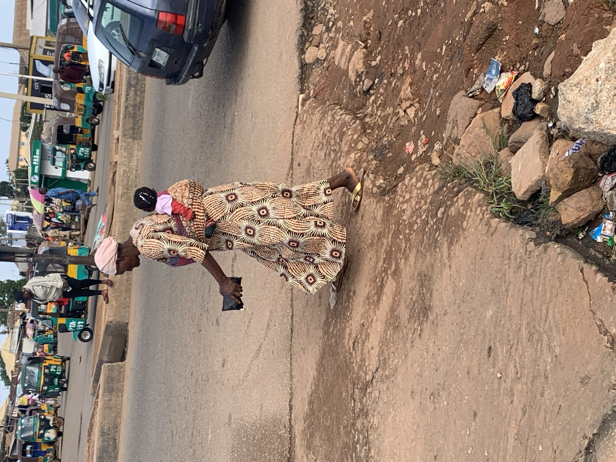 A woman wearing a patterned dress and headscarf walks with a baby strapped to her back, holding a black wallet in her right hand. The scene is set on a dusty, uneven road in Gura Top, Nigeria, with a backdrop of various auto-rickshaws and a dark car. The environment is characterized by rough terrain, scattered rocks, and a pile of discarded trash, including plastic bags and wrappers. Other individuals and vehicles are visible in the background, suggesting a busy urban setting.