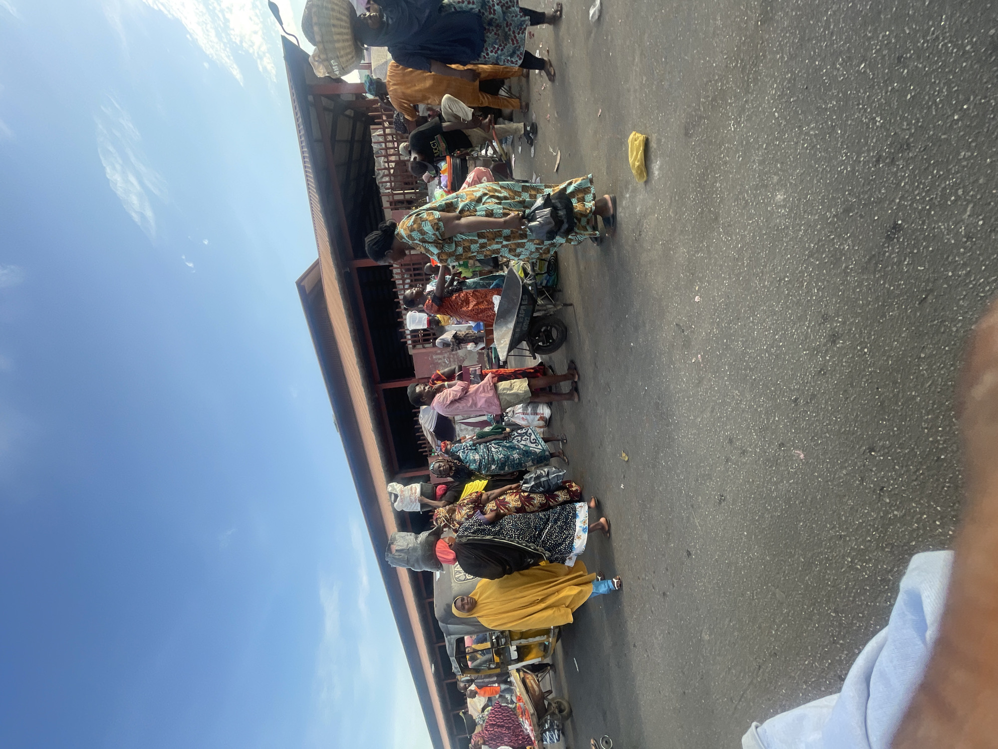 A street scene in Lokoja, Nigeria. Several people are gathered outside a building with a brown roof, walking, standing, and pushing carts laden with goods. Some people are carrying baskets on their heads. One individual is wearing a yellow garment, and another is in a patterned blue and brown dress. A yellow plastic bag lies on the ground. The sky is visible, with a clear blue hue. Part of a blue garment is visible in the bottom right corner of the image.