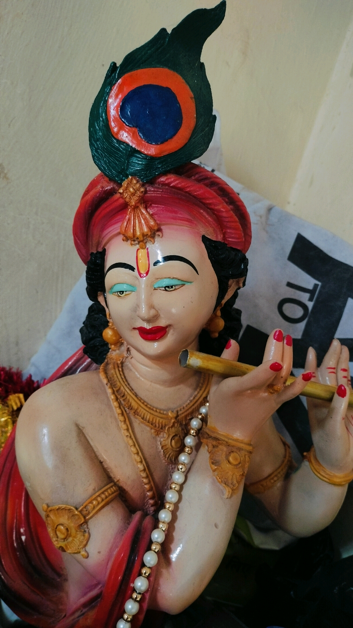 A close-up, eye-level, vertical shot features a colorful statue of the Hindu deity Krishna, prominently displaying a peacock feather atop his turban. The statue is crafted from a light-colored material, with intricate details and vibrant paintwork. Krishna is depicted with dark hair, a red turban adorned with a golden embellishment, and a peacock feather featuring shades of green, orange, and blue. His face is painted with wide, light blue eyes, defined eyebrows, and red lips. He wears a golden necklace, a pearl and gold bead necklace, and golden armbands. Krishna's hands are positioned around a golden flute, with red-painted fingernails. The background is a plain, light-colored wall. To the right of the statue, a portion of a white object with black lettering, possibly a box or packaging, is visible. The statue appears to be placed indoors, possibly in a home or temple setting. There is no discernible information within the image to determine its specific location within the city of Ramraj, India.