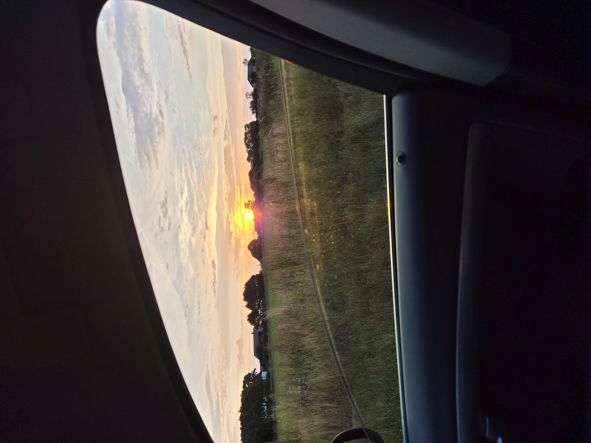 The image captures a serene sunset viewed from inside a vehicle, looking out through an open car window. The scene is set in the rural area of Gamla Köpstad, Sweden, as evidenced by the distant farmhouses, rolling fields, and distinct Scandinavian architecture visible. The sky is painted with hues of orange, pink, and yellow, with scattered clouds adding depth and texture to the sunset. The foreground is dominated by the interior of the car, with the dark grey car door panel taking up the bottom portion of the frame. The open window frames a landscape of tall, dry grass and scattered trees, with a few indistinct figures, possibly sheep, visible in the distance. The overall impression is one of peaceful contemplation of a rural Swedish sunset.