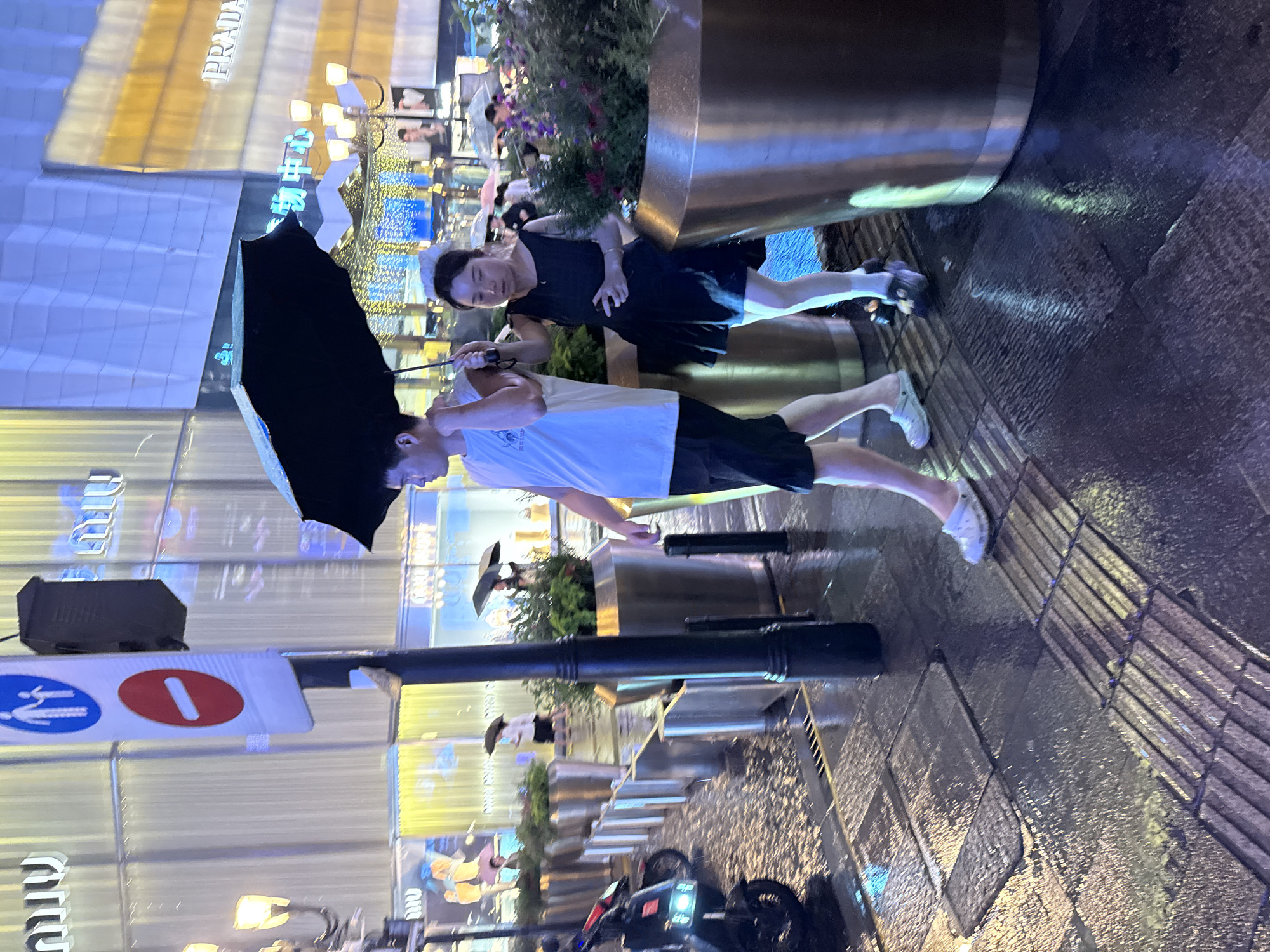 In Yuzhong District, China, a man and a woman walk under a black umbrella on a wet sidewalk at night. The man, wearing a white t-shirt and black shorts, walks slightly ahead of the woman, who is dressed in a black dress. The street is illuminated by streetlights and the neon signs of nearby businesses. The ground is wet and reflective, indicating it has been raining. Other people with umbrellas can be seen in the background, as well as planters with greenery, a scooter parked on the left, and a traffic signpost with pedestrian and no-entry symbols. The architecture of the buildings appears modern, with large glass facades and illuminated signage.