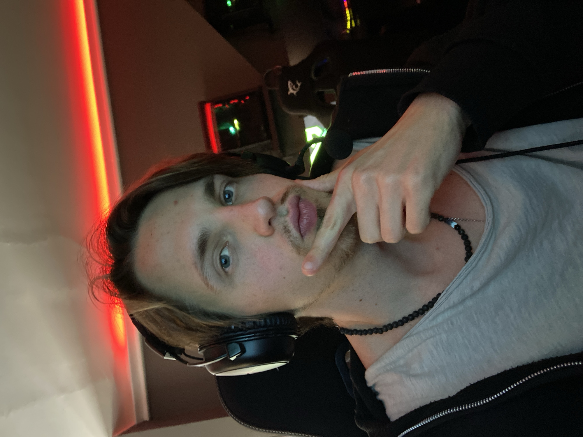 A young Caucasian male wearing a black hoodie, grey t-shirt, and black beaded necklace is pictured from the chest up. He is also wearing black headphones with white padding. He is looking directly at the camera with his right hand near his mouth. The background is dark, with red and green lighting elements suggesting a gaming setup. A gaming chair is visible. The location cannot be determined from this image. There is no information that the photograph was taken in the city of Bar, Montenegro.