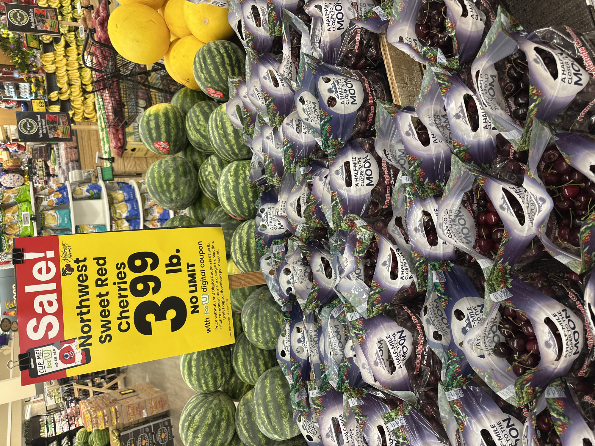 The image shows a display of fresh produce in a grocery store, likely a Mariano's or similar supermarket, in Schaumburg, Illinois, USA. In the foreground, numerous plastic bags of dark red cherries are stacked on wooden shelves. The bags feature branding with a moon and mountains, and text such as "A HALF-MILE CLOSER TO THE MOON" and "Premium High-Altitude Cherries." Behind the cherries, a large quantity of watermelons is visible. Further in the background, there are displays of other fruits and vegetables, including bananas and large yellow melons. A yellow "Sale!" sign is prominently displayed in the upper left quadrant of the image, advertising "Northwest Sweet Red Cherries" for "3.99 lb." with a "NO LIMIT" offer with a digital coupon. The store environment is illuminated by artificial lighting. There are no people visible in the image.