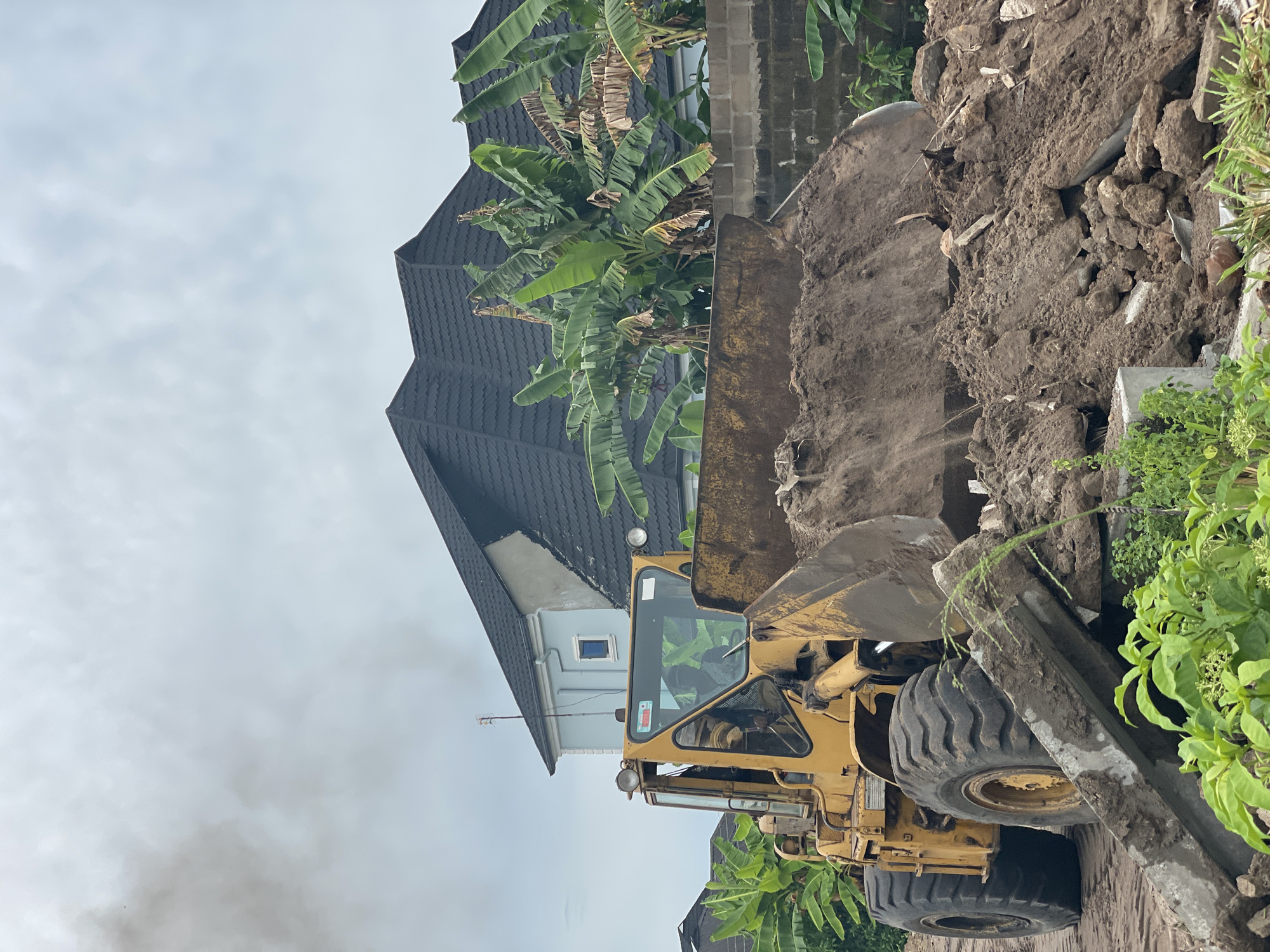 The image depicts a yellow front-end loader in Badagry, Nigeria, excavating a pile of dirt and debris. The loader is positioned close to a concrete barrier, with green vegetation growing nearby. In the background, there is a two-story building with a dark gray roof, and banana trees are visible. The sky is overcast.
