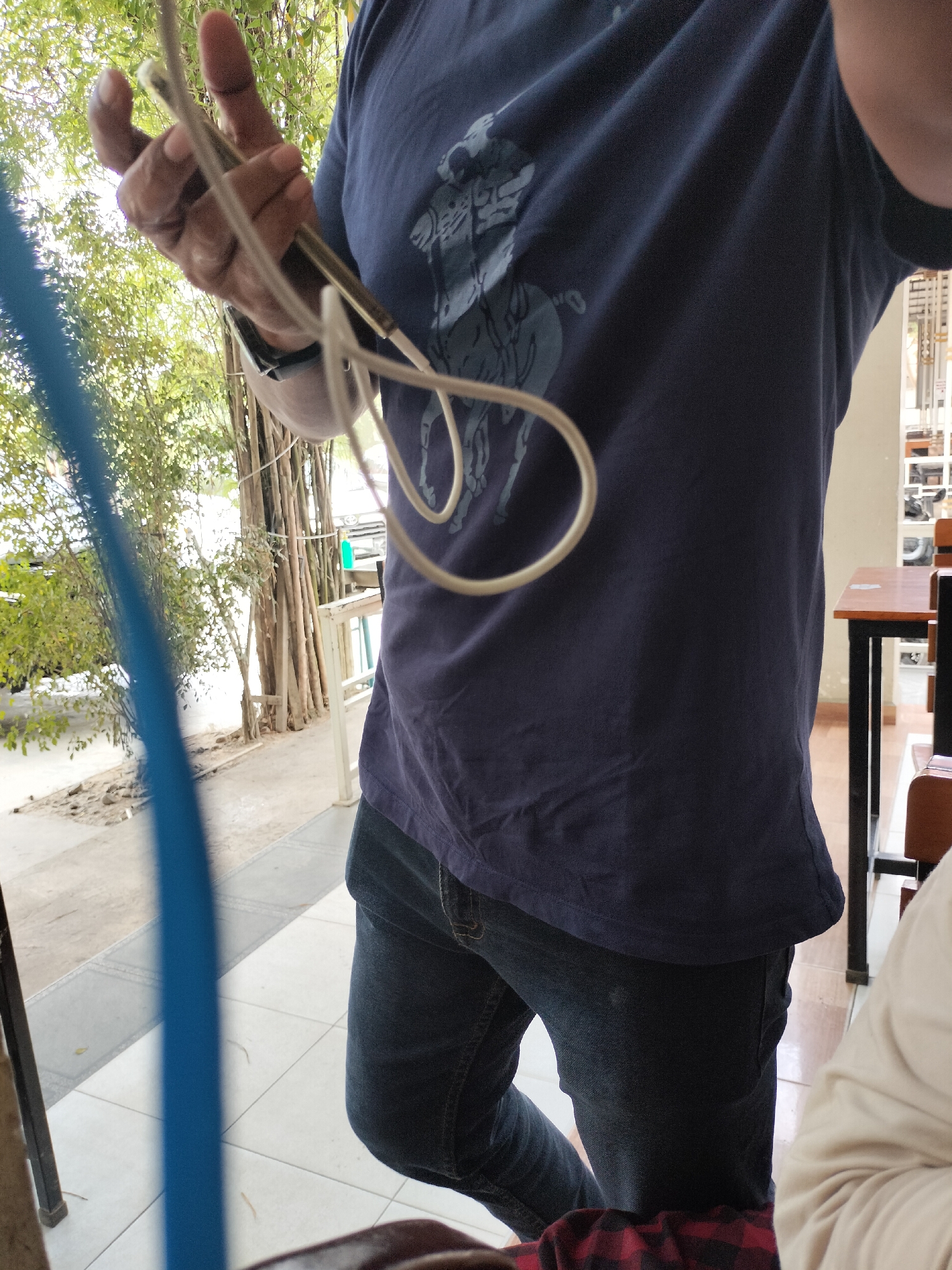 An individual wearing a dark blue t-shirt with a grey horse logo and dark wash jeans is holding a thin, light-colored cable or wire in their hands. The background suggests an outdoor or semi-outdoor setting with bamboo stalks and greenery visible. In the distance, there are tables and chairs, hinting at a cafe or seating area. The scene appears to be in Keudealue Ie Puteh, Indonesia.
