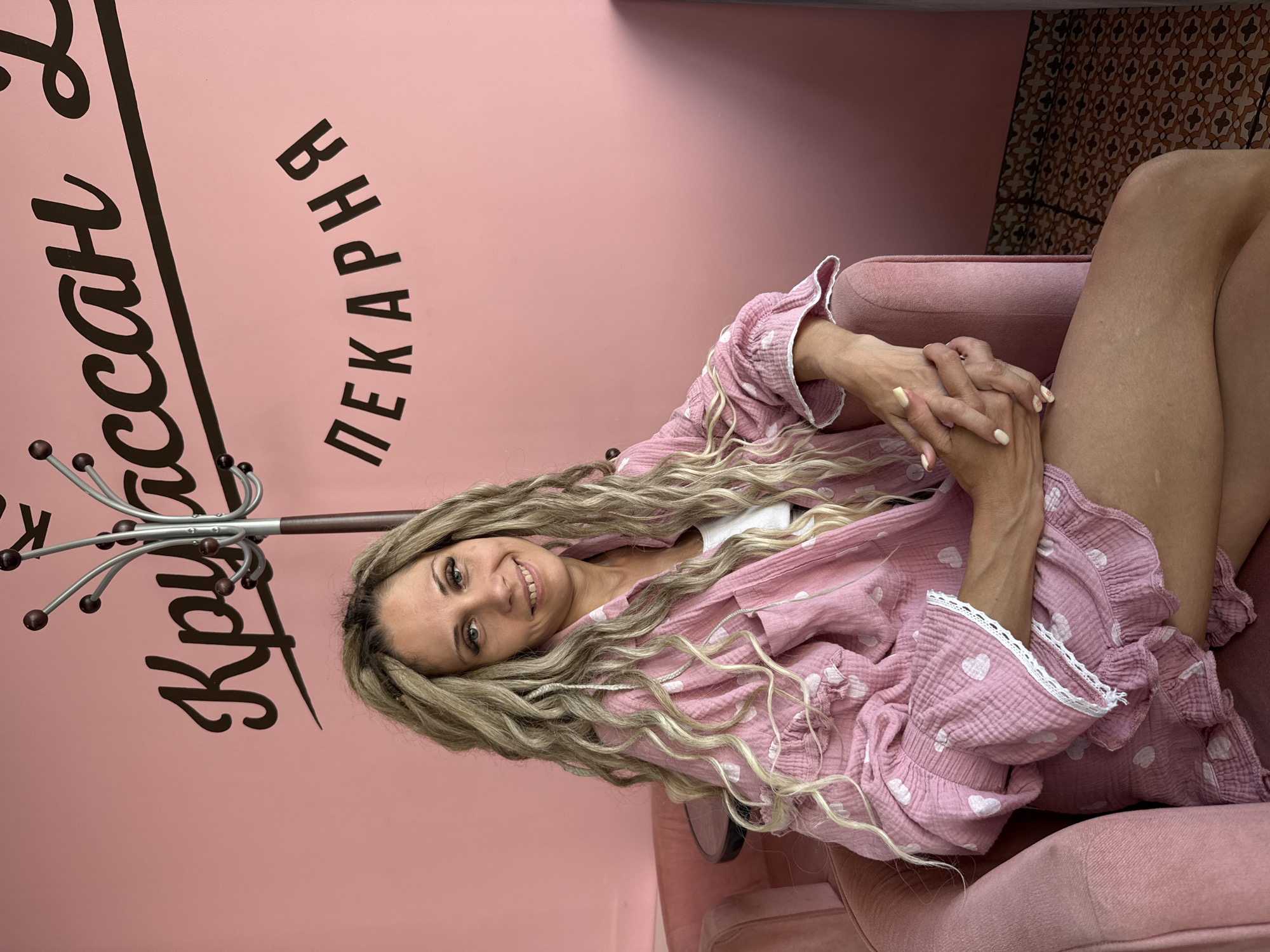 A woman with long, blonde dreadlocks is seated in a pink armchair against a pink wall. She is wearing a pink, heart-patterned pajama set. Her hands are clasped in her lap. Behind her, a coat rack stands in front of the wall, which features black text in a script font and block letters. The script text reads "Круассан" and the block letters read "ПЕКАРНЯ". The text translates to "Croissant Bakery". The setting is identified as a bakery in Rusanovo, Russia.