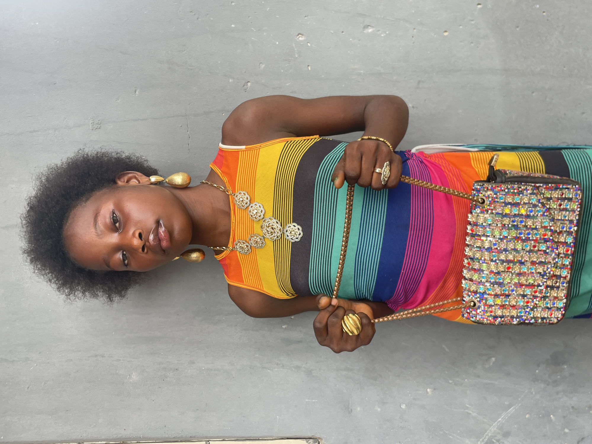 A young Black woman is depicted in a medium shot, lying on her back against a plain, light gray concrete surface, looking directly into the camera. Her dark, natural curly hair frames her face. She wears a vibrant, sleeveless dress or top featuring vertical rainbow stripes in shades of orange, yellow, green, blue, and pink.
Her accessories include prominent gold-toned jewelry: large teardrop earrings, a long necklace with multiple intricate floral medallions, a thin bracelet on her right wrist, and several rings, including a large, ornate one on her right hand and a textured one on her left. She holds a small, rectangular handbag with both hands. The bag is heavily embellished with multi-colored rhinestones or beads, giving it a sparkling appearance, and has a thin, braided gold-toned strap. Her expression is neutral to slightly serious. The setting appears to be outdoors or in a simple concrete space, typical of locations in Badagry, Nigeria. There is no visible text.