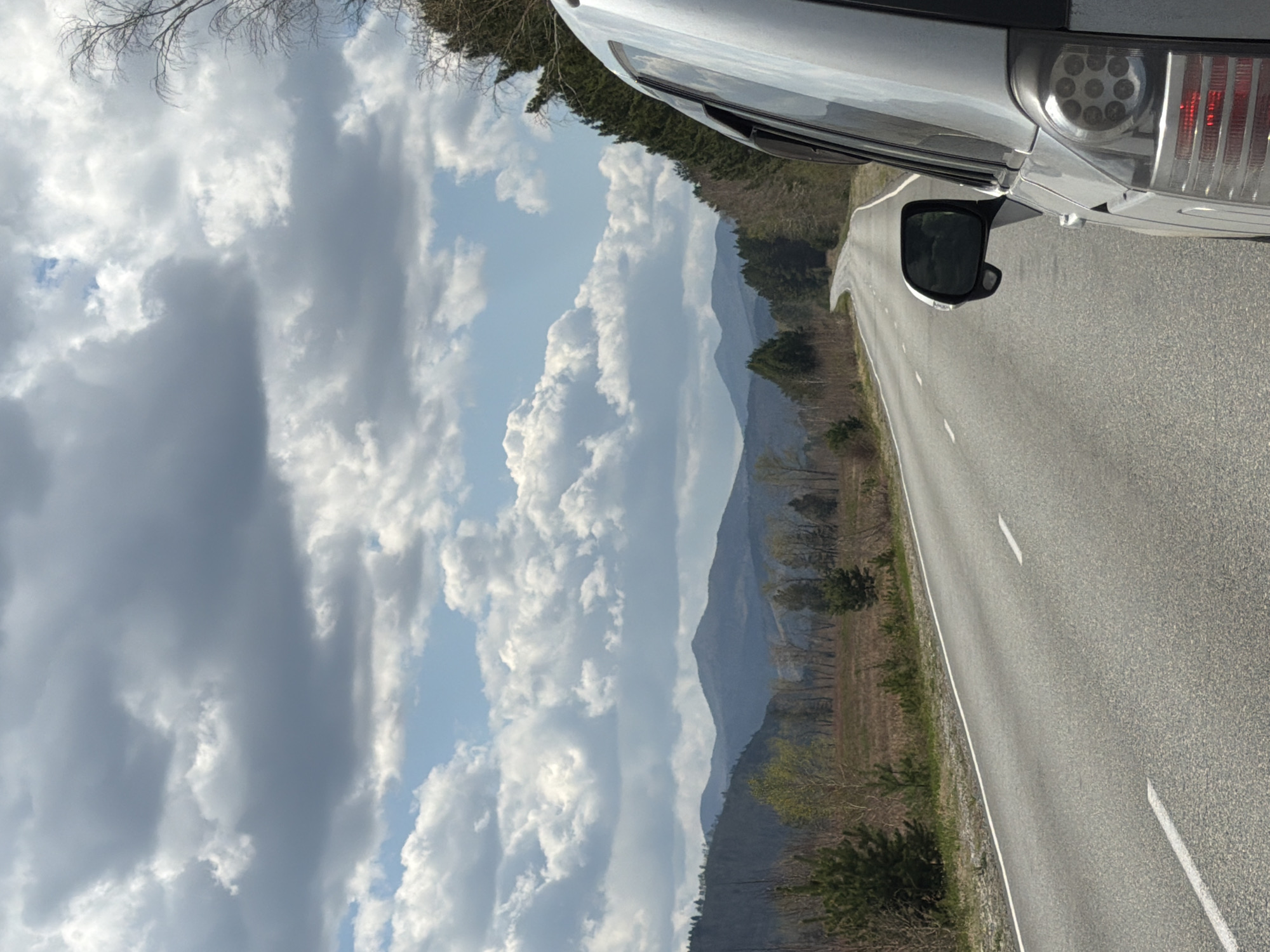 The image shows a section of asphalt road with white lane markings, likely taken from inside a vehicle. The right side of the vehicle's front portion, including the side mirror and headlight, is visible in the upper right. The road leads towards distant, hazy mountains. A line of trees runs along the edge of the road. The sky is filled with dramatic, gray and white clouds. The location may be Сизая, Russia, as specified.