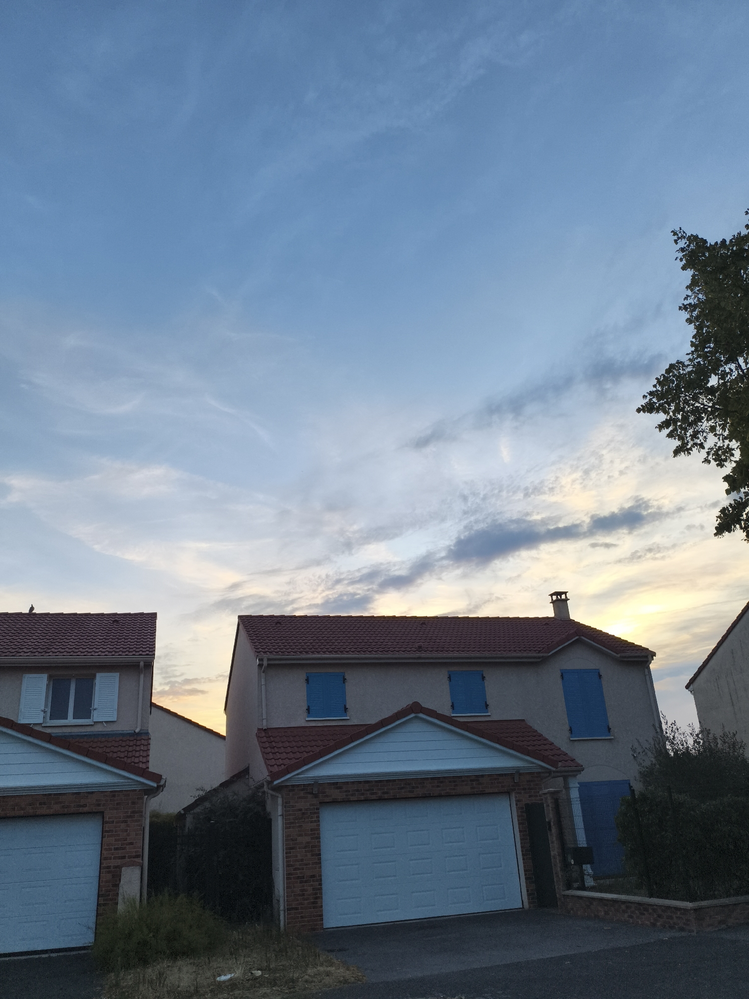 In Carrières-sous-Poissy, France, two attached houses with red tile roofs and light-colored facades are visible at dusk. The house on the left has a white garage door and a window with white shutters. The house on the right features a red brick facade on its garage, a white garage door, and two windows with bright blue shutters. A narrow gap between the two houses reveals overgrown vegetation and a portion of another building. The sky above is a mix of pale blue and wispy clouds, with the setting sun casting a warm glow on the horizon. A tree branch with dark green leaves extends into the frame from the upper right corner.