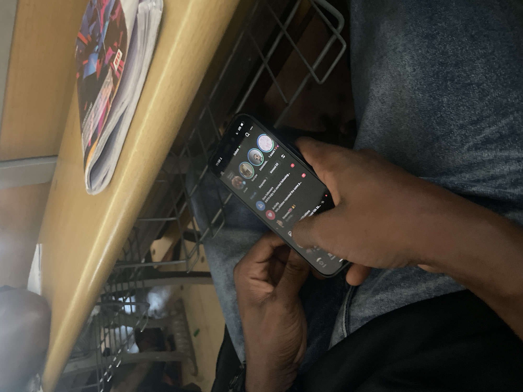 A person's hands are shown holding a smartphone, displaying an application with multiple circular profile pictures and text messages. The phone is held over a lap covered in blue denim fabric. In the background, a wooden surface, possibly a desk or table, is visible, with a stack of papers or a magazine resting on it. Below the wooden surface, a metal shelf structure is partially visible. The overall environment suggests an indoor setting, potentially a classroom or study area, located in Ihiagwa, Nigeria.