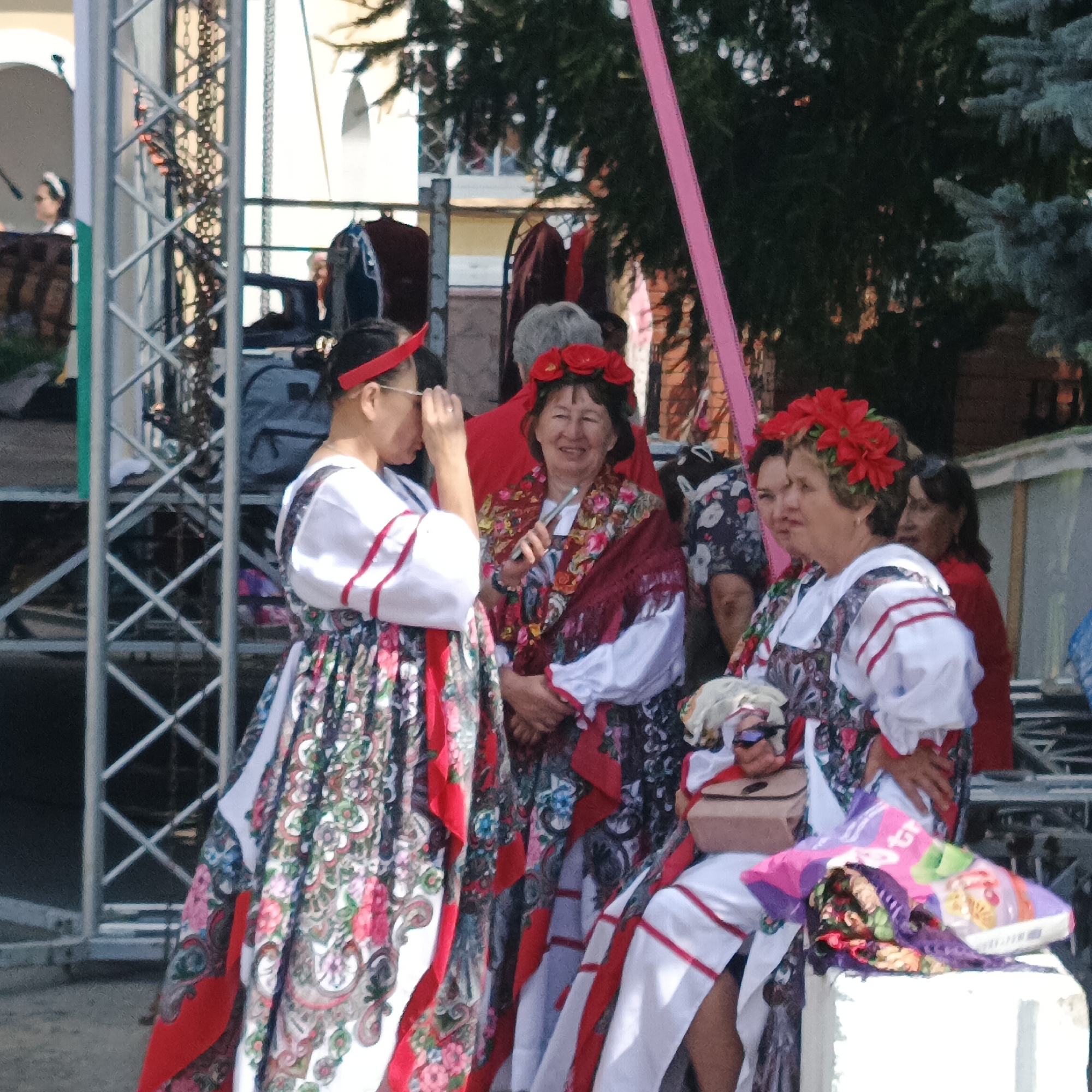 In Nikolo-Beryozovka, Russia, a group of women dressed in traditional folk costumes are gathered outdoors. The costumes feature white blouses with red trim and elaborately patterned skirts and scarves in shades of red, white, and green. Red floral headbands adorn the heads of some women, and one woman has a red flower crown. They appear to be standing near a stage or event setup, with metal scaffolding visible in the background. The women are interacting, with some looking at each other and one holding a small object. The overall atmosphere suggests a cultural event or festival.