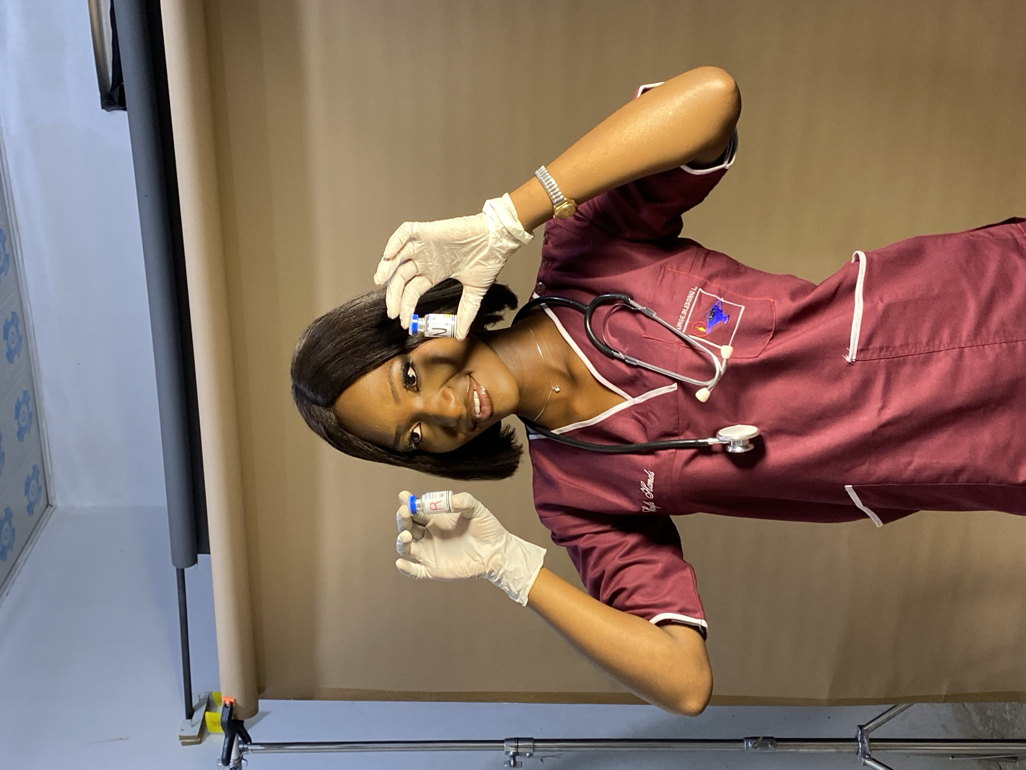 A young, Black woman in a burgundy medical uniform with white trim poses indoors with a stethoscope around her neck and white latex gloves. She is holding two small vials with blue and white labels. The woman is wearing a watch on her left wrist and a necklace. Behind her is a seamless paper backdrop of tan and black, set up with silver metal stands and clamps on a white floor. The room has a section of blue and white patterned wall paneling on the left edge. No clear indicators suggest this photo was taken in Gura Top, Nigeria.