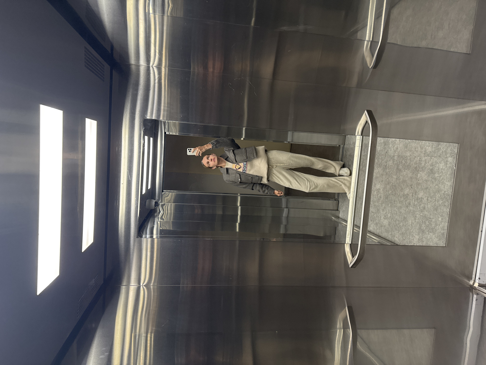 A photograph taken inside an elevator shows a woman holding a smartphone and taking a selfie. She is wearing a gray jacket and light-colored pants. The elevator's interior is predominantly stainless steel with a textured floor. Bright light is visible from the elevator's ceiling lights. The location is unknown.