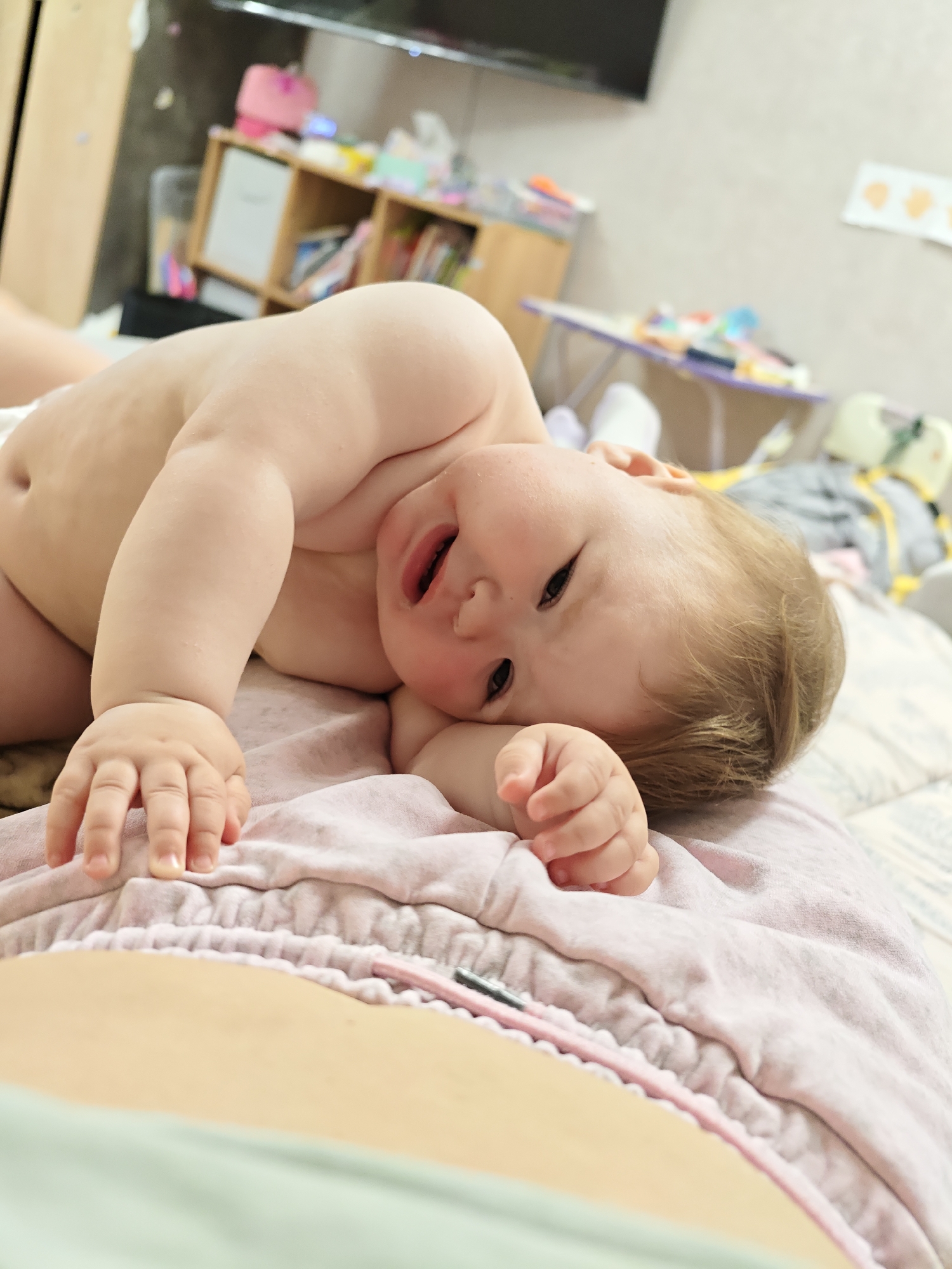 An intimate, close-up portrait captures a baby lying on its side, looking directly at the viewer with a slightly open mouth and an ambiguous expression that could be inquisitive or on the verge of whining. The baby, appearing to be a toddler, is topless, wearing only a white diaper, and has light brown hair. Its left arm is extended towards the foreground, hand open, while its right arm is tucked beneath its head.
The baby rests on a soft, pink fabric, possibly a blanket or garment, with a visible drawstring or elastic band. In the immediate foreground, a portion of an adult's torso or lap is visible, clad in light green or grey fabric, suggesting the baby is lying on or near the adult.
The background depicts a domestic indoor setting, likely a bedroom or living area in Chaikovsky, Russia. To the left, a light wooden shelving unit or cabinet holds various household items, including books and pink objects. A dark flat-screen television is mounted on the light-colored wall above it. Further in the blurred background, a small table with items and a yellow and grey baby accessory (possibly a bouncer or playpen part) are visible. The scene is well-lit, suggesting either daytime indoors or artificial lighting. There is no visible text in the image.