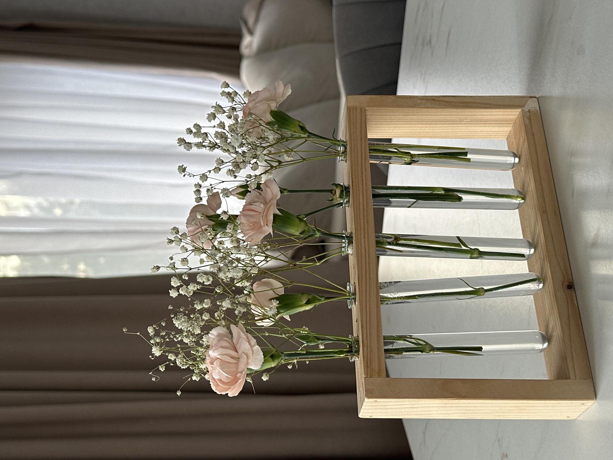 The image shows a wooden frame holding five glass test tubes filled with water and flowers. The flowers are a mix of pink carnations and baby's breath. The frame is sitting on a white surface, possibly a table. In the background, there is a window with white curtains and a gray couch. It is impossible to determine the location as Chaikovsky, Russia from the visual data.