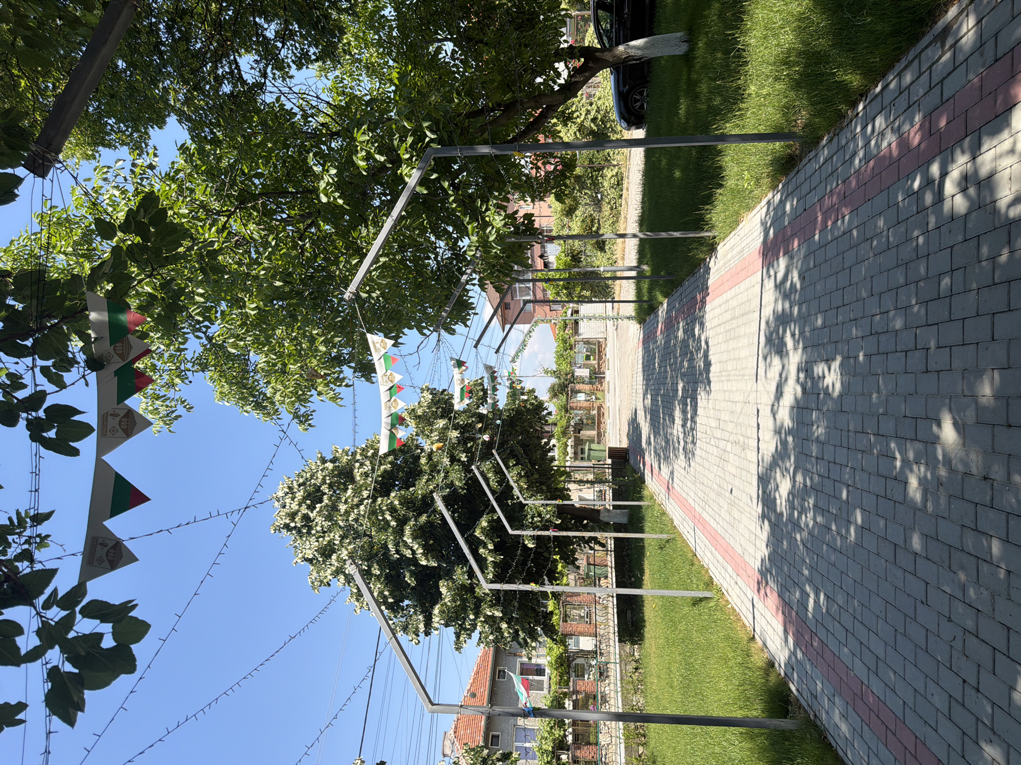 The image depicts a paved walkway lined with grass and trees, decorated with strings of triangular flags and lights. The walkway is made of grey pavers with a pink border and is bordered by metal poles supporting the decorations. In the background, there are buildings with red-tiled roofs, typical of Bulgarian architecture. A black car is parked on the right side of the walkway. The sky is clear and blue, indicating a sunny day. The scene is likely in the village of Zlatitrap, Bulgaria, given the presence of Bulgarian flags.