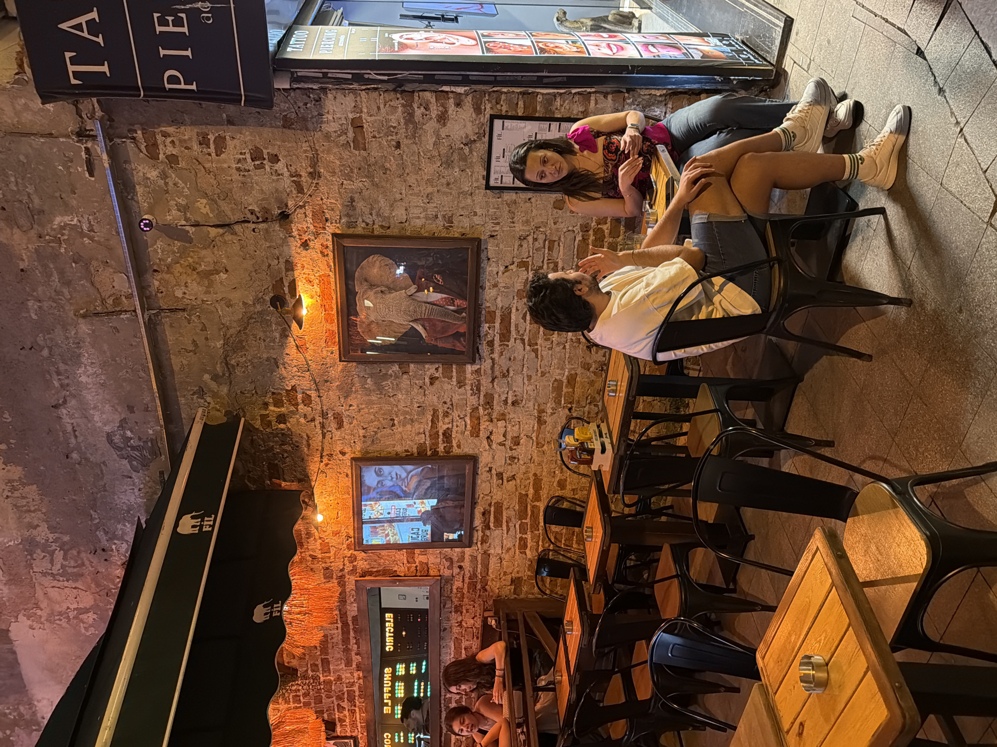 The image depicts an outdoor seating area of a establishment, likely a cafe or restaurant, with exposed brick walls and a somewhat rustic ambiance. In the foreground, a man and a woman are seated at a table. The man, wearing a white t-shirt and denim shorts, is gesturing with his right hand as he speaks. The woman, in a patterned top and jeans, is looking towards him, holding a cell phone in her left hand and a white object, possibly a packet of sugar or a napkin, in her right. There are two ashtrays on their table.
The seating area features metal chairs with wooden seats and several wooden tables. In the background, another pair of women are seated at a table, with one looking at a menu. A neon sign displaying "ELECTRIC SHUFFLE" is visible reflected in a mirror on the wall behind them. Artwork, including a framed picture of an elephant in a suit, adorns the brick wall. A sign with "FIL" and elephant logos is visible above the seating area, suggesting it is the establishment's branding. To the right, a sign for "TATTOO PIERCING" indicates a neighboring business, and a statue of a dog is partially visible. The overall lighting is dim and warm, suggesting an evening scene. The location is identified as Kadıköy, Turkey.