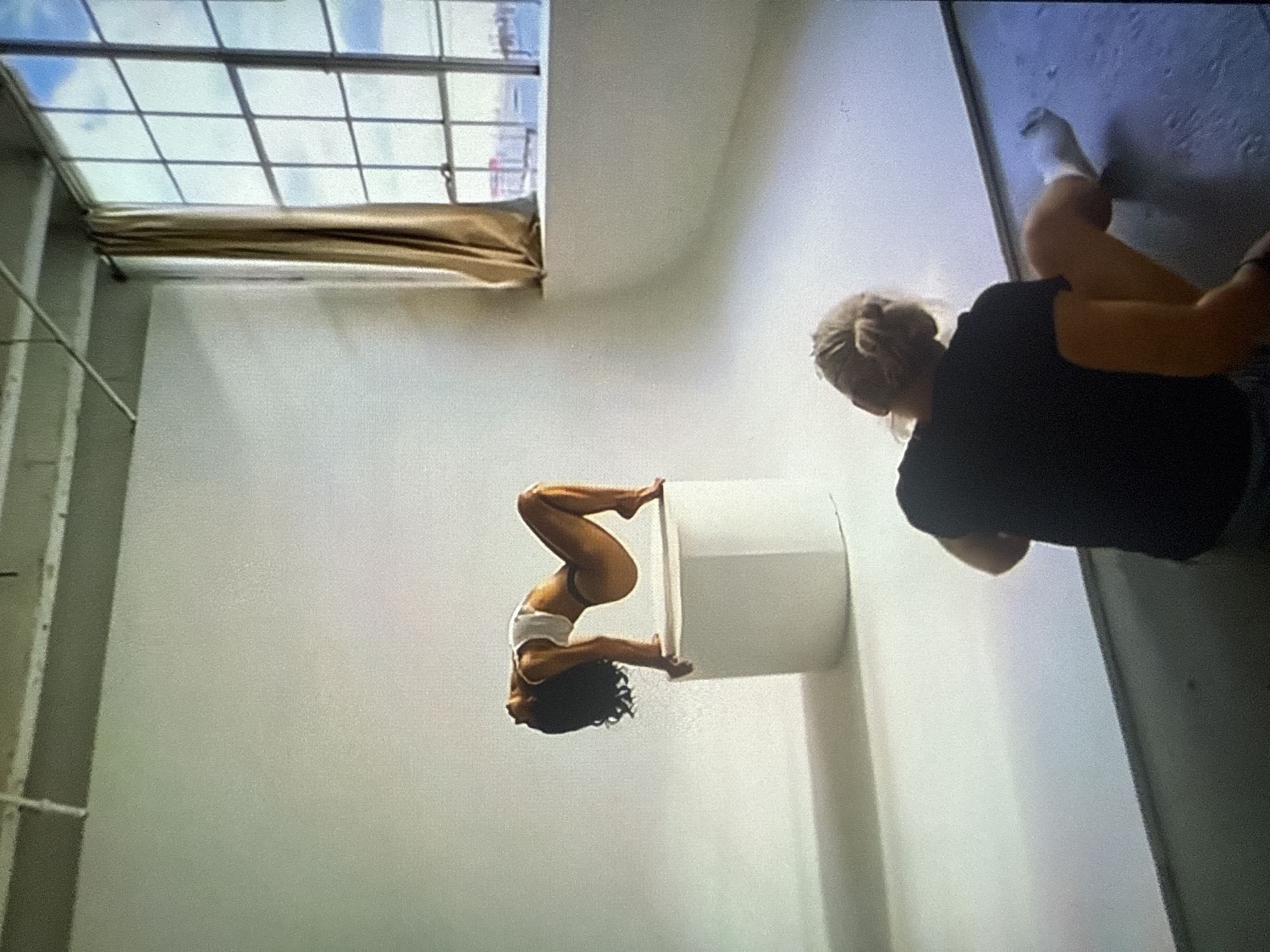 The image depicts an indoor photography studio during a photoshoot. In the midground, a female model with dark, curly hair, wearing a white crop top and dark briefs, is performing a flexible pose on a white cylindrical pedestal. Her back is arched, head tilted backward, with her hands and feet supporting her on the edges of the pedestal. The background behind the model is a continuous white cyclorama, extending from the floor to the wall.
To the right, a large, multi-paned window reveals an outdoor urban scene under a partly cloudy sky. Through the window, a body of water is visible, with distant buildings and what appears to be part of a port or industrial area, including a red structure. A light-colored, possibly beige, curtain hangs to the left of the window frame. The studio ceiling features exposed concrete beams and conduits, characteristic of an industrial or loft space.
In the foreground, with her back to the viewer, a person with blonde hair tied in a bun, wearing a black short-sleeved shirt and dark bottoms, is seated on a darker gray floor area, looking towards the model. Her right leg is bent, and she wears a white sock on her right foot. This individual appears to be a photographer or a member of the production crew, observing the model.
The image itself does not contain any specific identifying markers or landmarks to confirm the location as Varna, Bulgaria. The visible cityscape through the window is generic and could represent various waterfront urban environments.