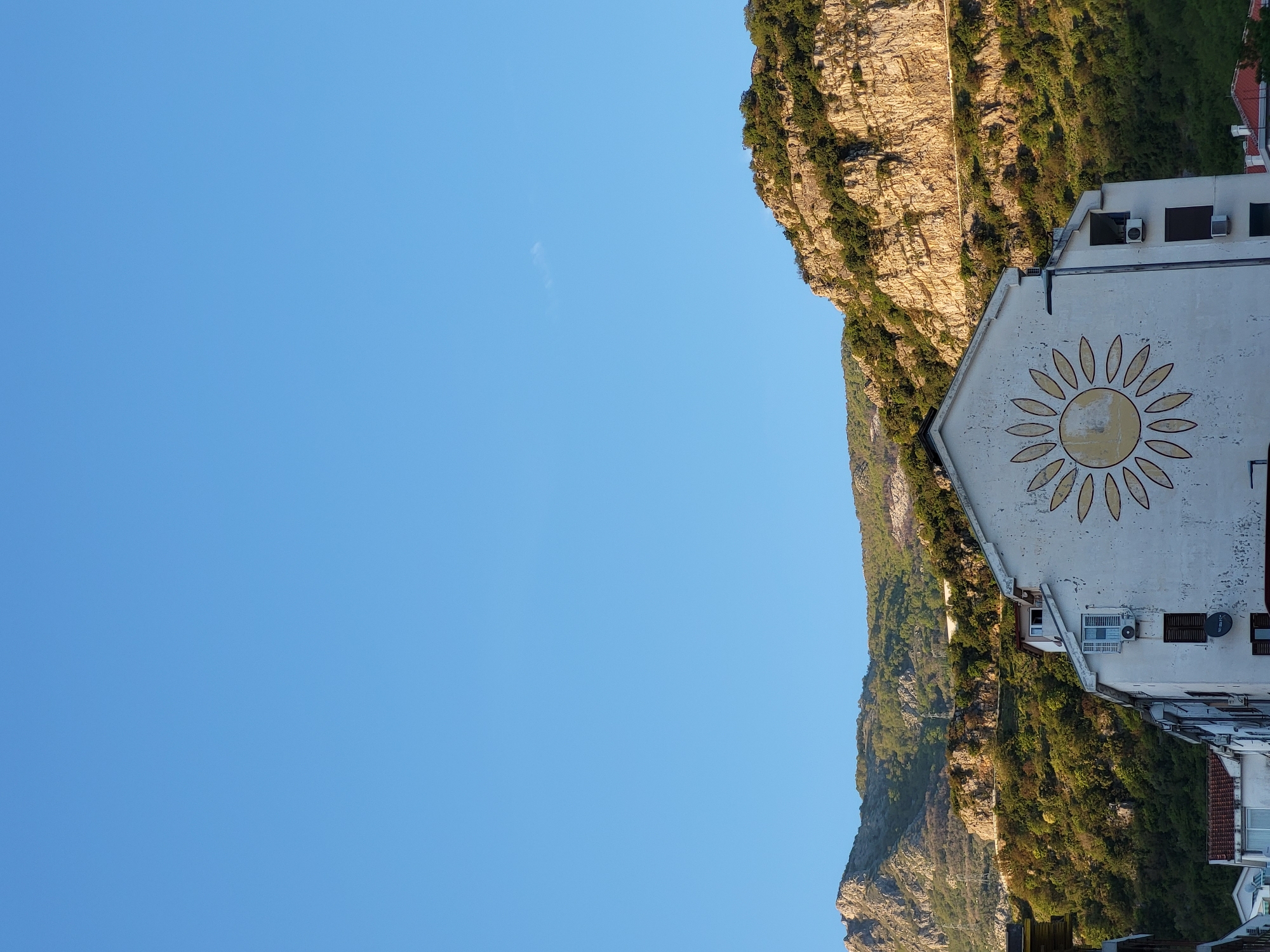 The image, when viewed in its correct upright orientation, captures a bright, sunny day in Budva, Montenegro, showcasing a striking blend of urban architecture and natural landscape.
On the right side of the frame, a light-colored building, possibly white or off-white, stands out with a large, decorative sunflower painted prominently on its upper facade. The sunflower design features a yellow center and orange petals. The building has visible architectural details including windows, some equipped with dark shutters, and outdoor air conditioning units. A satellite dish is also affixed to its side. A glimpse of another building with a terracotta-tiled roof can be seen partially at the bottom right.
The left and central background is dominated by a majestic, rugged mountain. Its slopes are richly covered in green vegetation, interspersed with large, exposed sections of rocky terrain that catch the sunlight, creating varied textures and shadows. The mountain stretches across the midground, forming a natural backdrop to the building.
Above the mountain and to the left of the building, a vast expanse of clear, bright blue sky fills the upper portion of the image. A single, very faint, wispy cloud is barely discernible in the expansive blue. The overall scene exudes a peaceful and warm atmosphere, characteristic of a coastal Mediterranean setting under clear weather conditions. No people or specific activities are depicted, and no visible text is present.