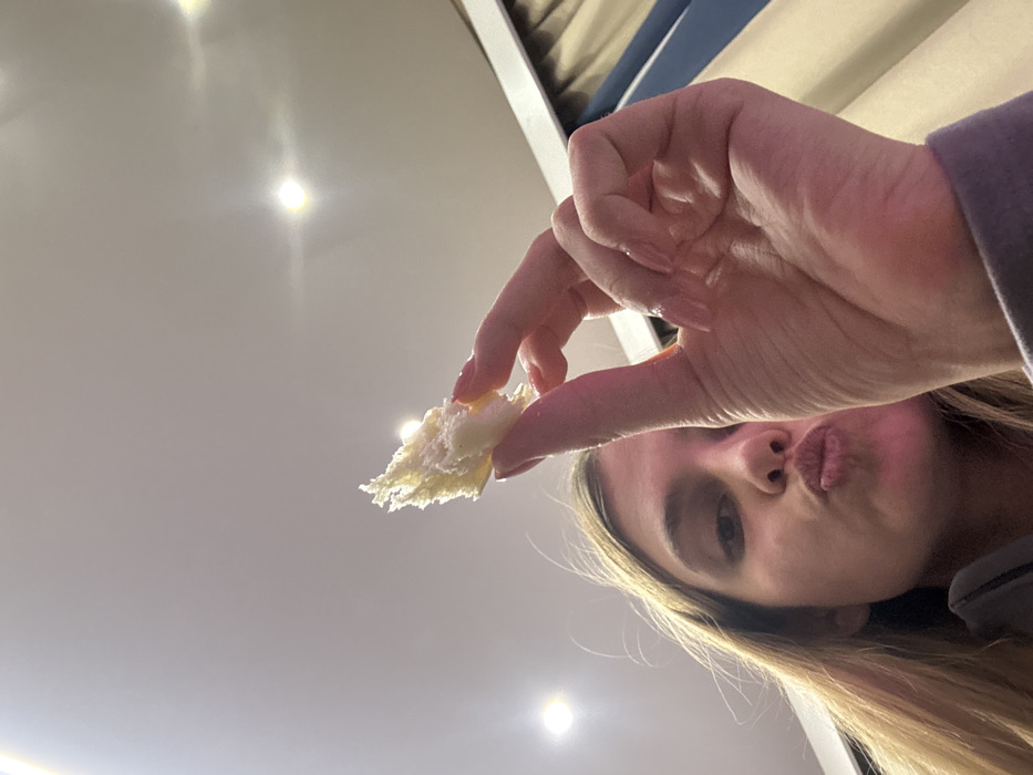 A woman holds a piece of bread in her fingers, positioned between the camera and her face. Her lips are puckered in a kiss expression. She has fair skin and long blonde hair. Overhead, bright recessed lighting illuminates a white ceiling. A hint of a bunk bed structure is visible in the background.