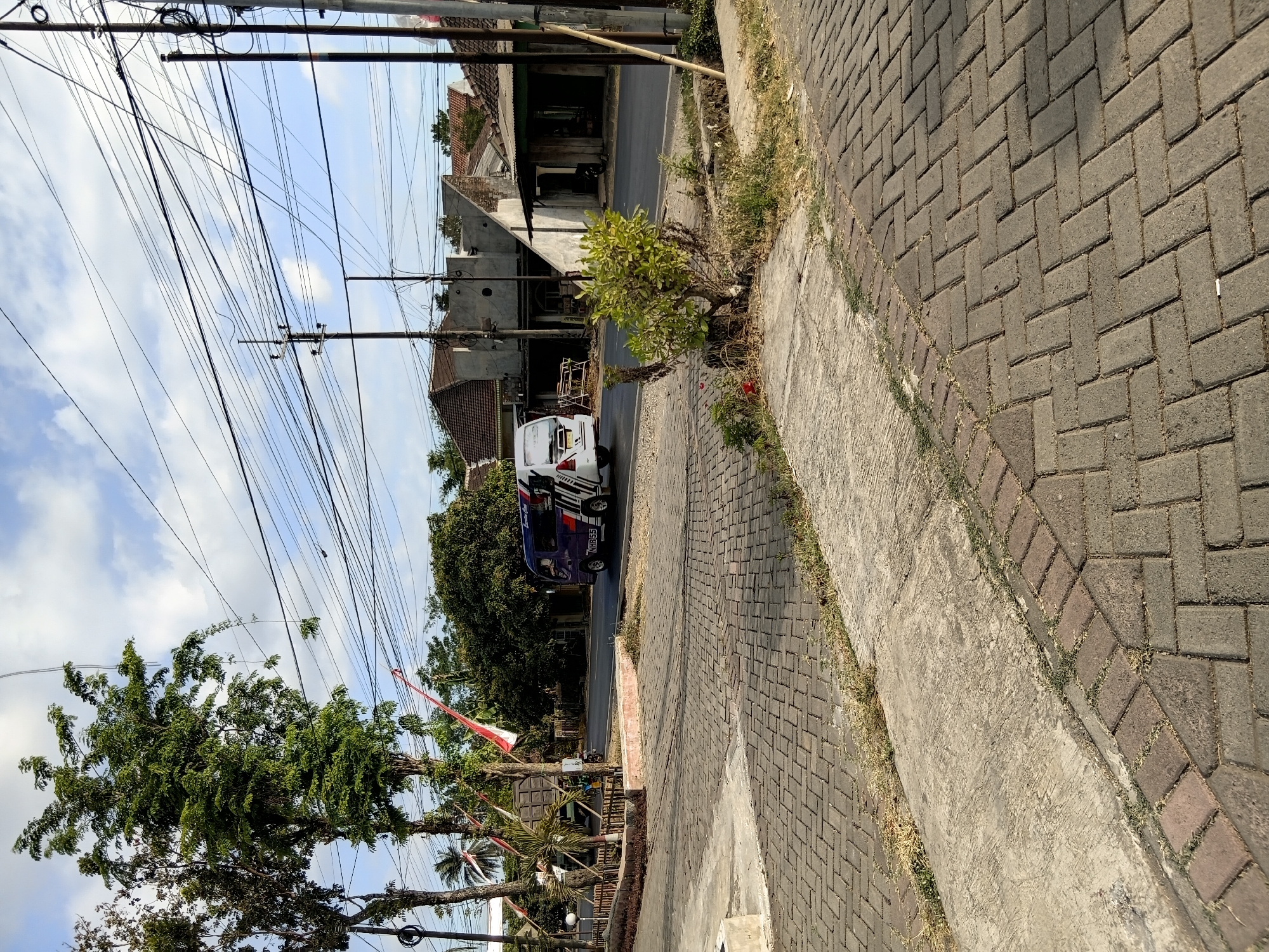 The image captures a street scene in Tanggul Wetan, Indonesia, under a partially cloudy sky. A purple and white van, identified by the license plate "NMB 55", is traveling on the asphalt road. To the left of the road, a brick walkway leads towards a low wooden fence bordering a paved area. Behind the fence, trees and some red and white Indonesian flags are visible. Across the road, several buildings with tiled roofs are situated, some appearing to be shops or residences. Overhead, a dense network of power lines crisscrosses the sky, supported by utility poles. The foreground is dominated by a paved pathway made of rectangular bricks. A small bush with green foliage is growing in the middle ground, near the roadside. The overall impression is that of a typical street in a residential or commercial area within an Indonesian town.