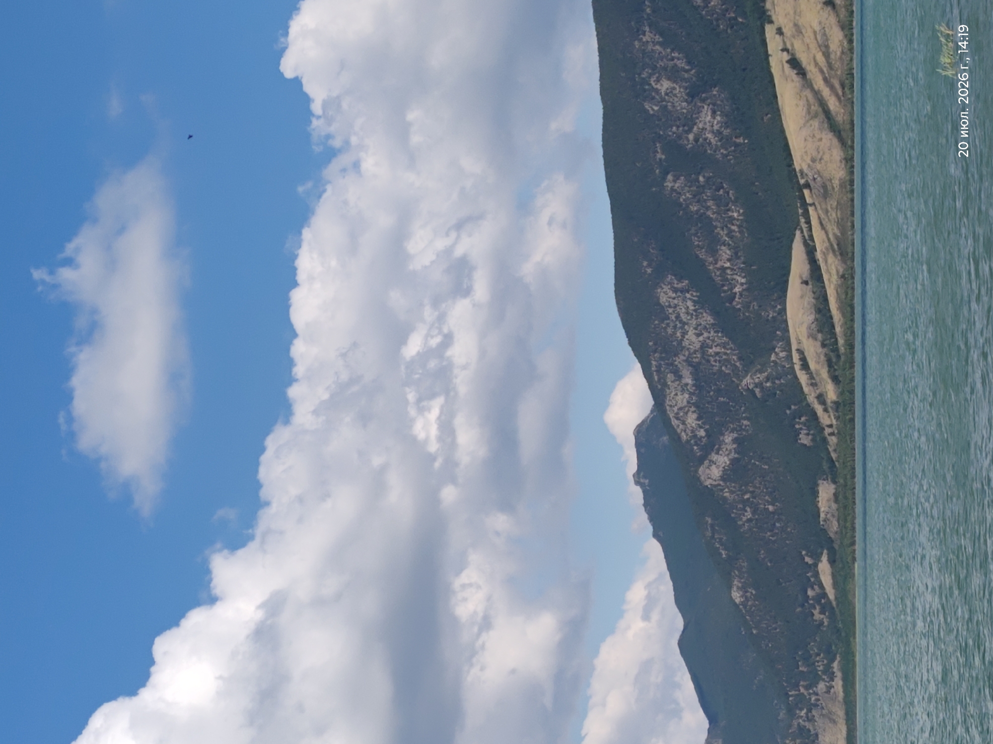 The image captures a serene natural landscape with a body of water in the foreground and a mountain range in the background under a partly cloudy blue sky. The water is a tranquil teal color, reflecting the sky. The mountains are covered in dense green vegetation and rocky outcrops, with a lighter, possibly grassy, slope leading down towards the water. A small dark shape, possibly a bird, is visible in the sky. A timestamp in the lower right corner indicates "20 июля 2026 г., 14:19", which translates to "July 20, 2026, 14:19". The location is specified as Акылбай, Kazakhstan.