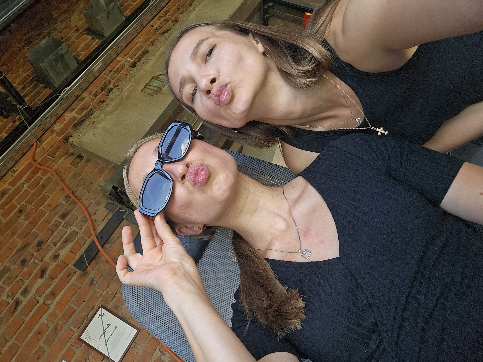 Two young women are taking a selfie outdoors in front of a brick wall. The woman on the left is wearing black, sunglasses, and a silver necklace with a square pendant. She is holding up her sunglasses with her right hand and puckering her lips. The woman on the right is wearing a black tank top and two gold necklaces, one with a heart pendant and another with a cross pendant. She is also puckering her lips. The background shows a red brick wall and some industrial-looking structures. A sign with Cyrillic text is visible on the wall to the left, which reads "ПОРТ". This suggests the location is in Chaikovsky, Russia, as the city is known for its port facilities.