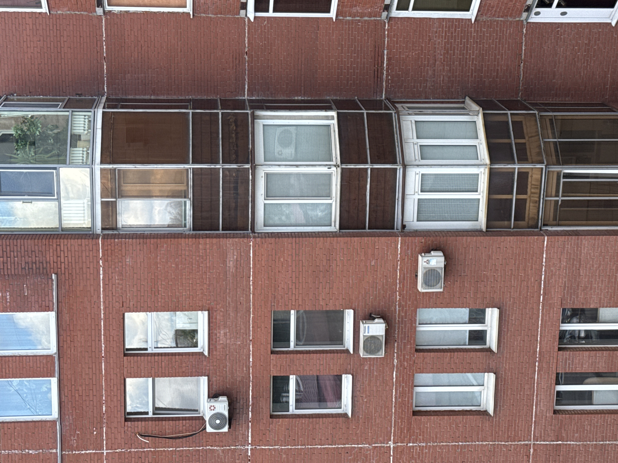 This is a high-angle, medium shot of a red brick apartment building, likely located in the city of Perm, Russia. The building features multiple windows, many of which are rectangular and have white frames. Several windows have blinds or curtains drawn, obscuring the interior.
Prominently visible are three air conditioning units attached to the exterior wall. Two are situated on the lower half of the building, and one is on the upper half. The building also has a multi-story bay window section with a hexagonal shape. This section features numerous windows with white frames and dark brown paneling.
The brickwork is laid in a horizontal pattern, with thin white lines running horizontally between some rows of bricks, possibly indicating structural seams or decorative elements. The sky is visible in some of the windows, reflecting clouds. No people are visible in the frame, and the scene depicts a static architectural exterior.