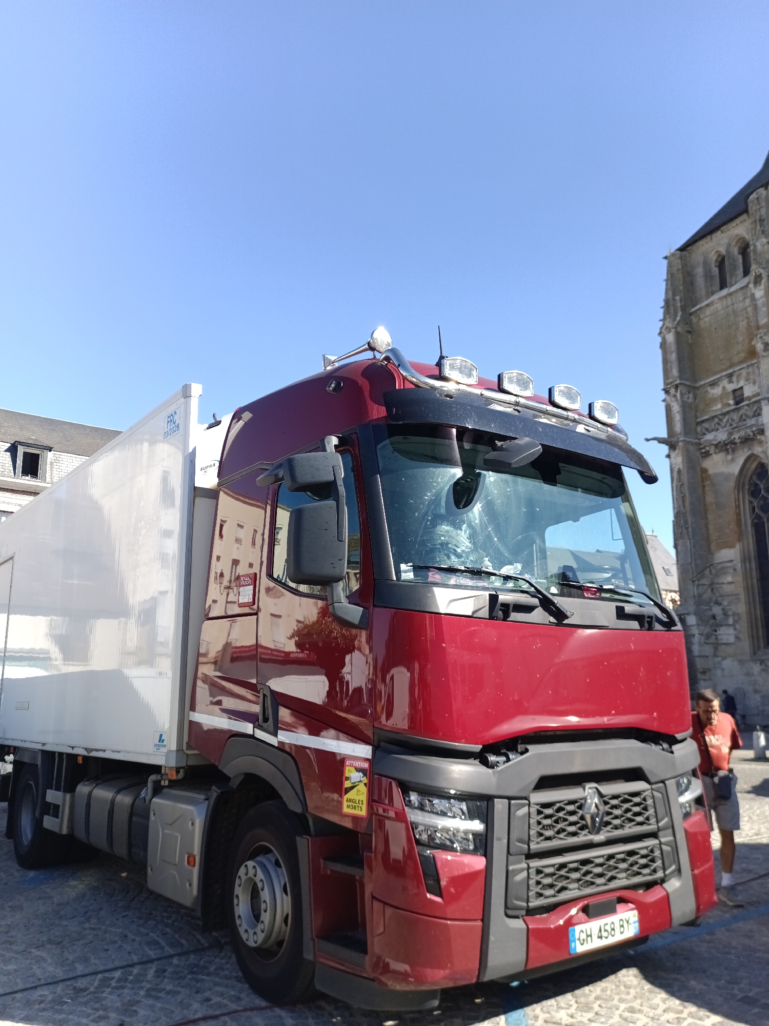 The image shows a red semi-truck parked on a cobblestone street in front of a stone building with arched windows, likely a church, in Le Neubourg, France. The truck has a white refrigerated trailer attached. A man wearing a red shirt and grey shorts is standing next to the truck, partially visible. The sky is clear and blue.
