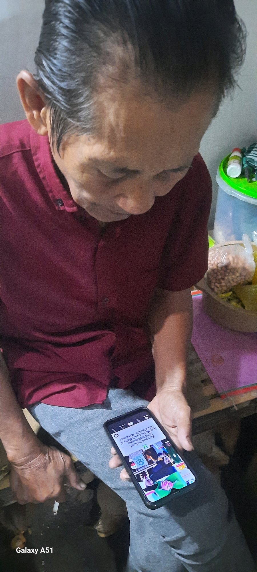 A man is seated, looking down at a smartphone in his hands. He is wearing a maroon short-sleeved shirt and grey pants. The phone displays an article with Indonesian text and an image of people in what appears to be an event. The background is blurred but shows a plastic container with items inside, suggesting a domestic setting. The lower left corner of the image displays "Galaxy A51" branding, indicating the smartphone's model. The scene is consistent with a person using their phone in Selopuro, Indonesia, as part of their daily life.