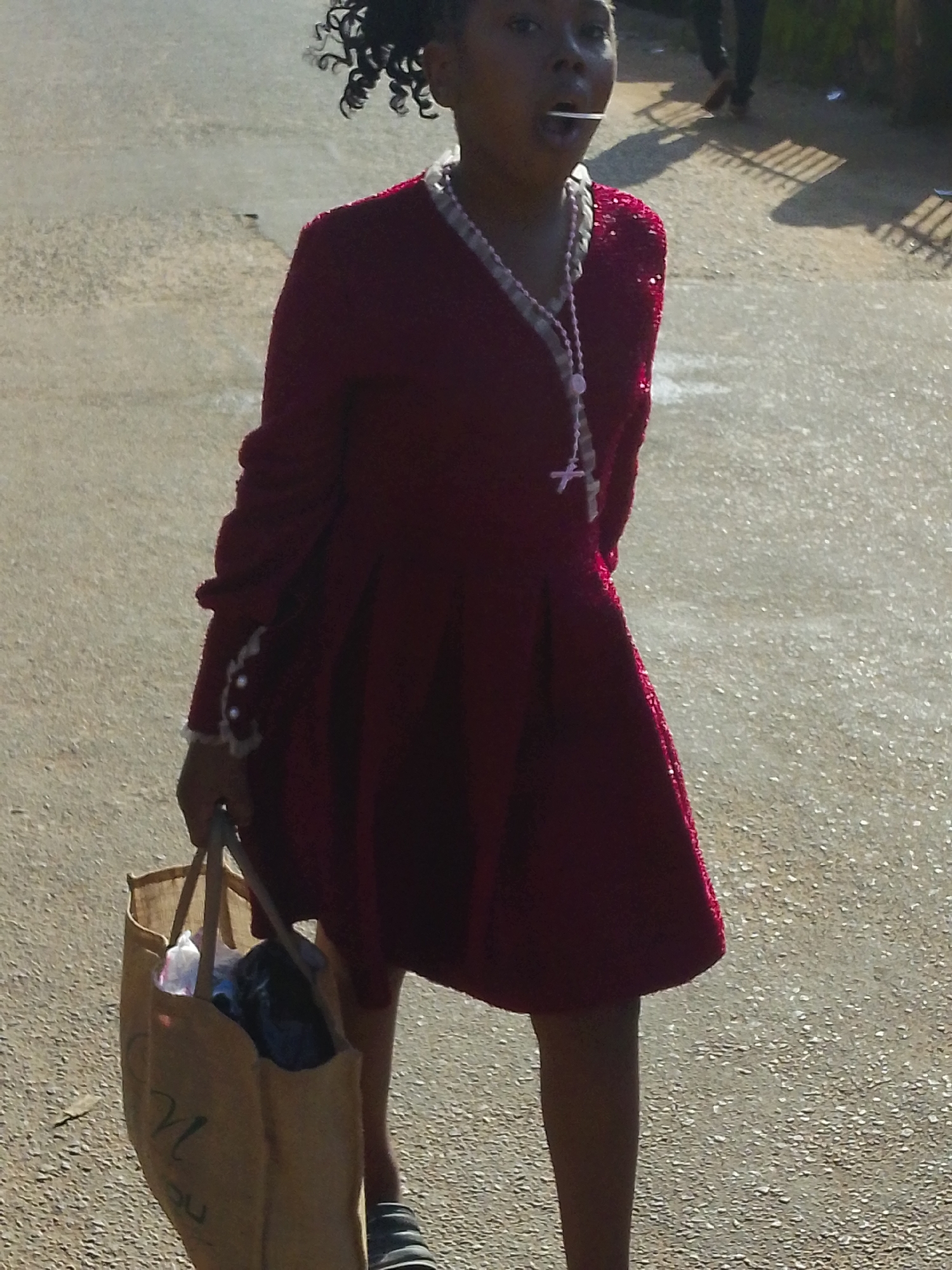 A young Black girl wearing a sparkly maroon dress and a rosary necklace walks outdoors on a paved surface, carrying a beige canvas bag filled with items. She has dark, curly hair and is holding a white lollipop in her mouth. In the background, a person wearing dark clothing walks away. The scene appears to be a street in Jos, Nigeria, suggested by the architecture and ambiance, though not explicitly identified. The lighting suggests it is daytime, possibly late afternoon due to the long shadows cast.