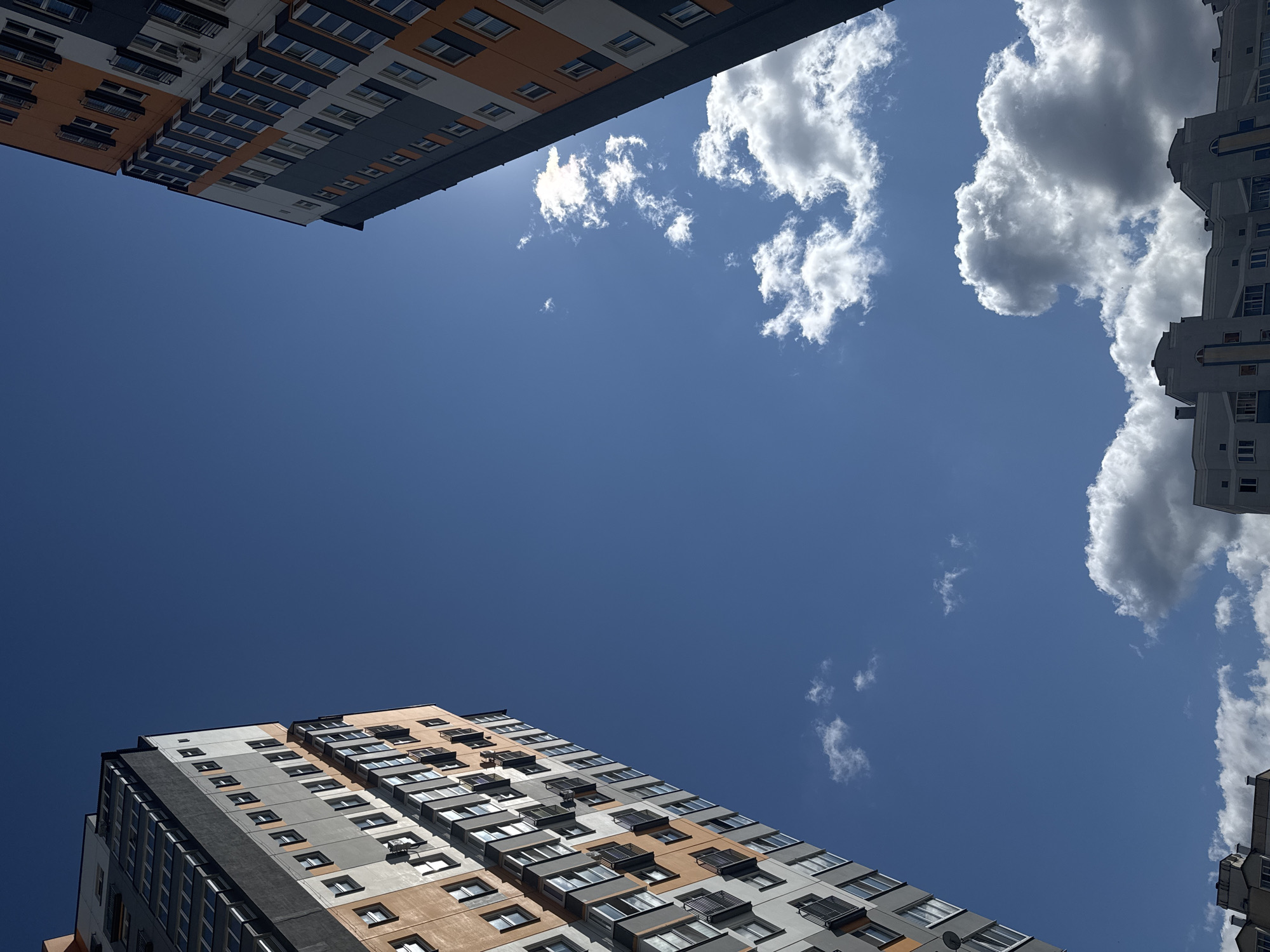 The image captures a low-angle view of two multi-story apartment buildings against a bright blue sky with scattered white clouds. The buildings, with their distinct modern architectural style featuring alternating segments of grey, white, and orange facades, are positioned on the left and right sides of the frame, creating a sense of depth. The left building exhibits a pattern of regularly spaced windows and balconies, while the right building displays a similar structure with a darker grey exterior dominating its visible sections. Sunlight subtly illuminates parts of the buildings and clouds, casting soft shadows. The environment appears to be an urban residential area within the Бараўлянскі сельскі Савет, Belarus, suggesting a contemporary housing development. No people or specific actions are depicted, focusing the composition on the architectural elements and the expansive sky.