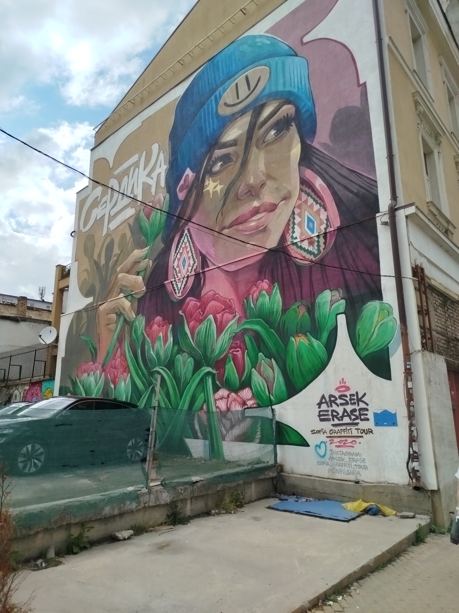 A large and vibrant mural depicting a stylized woman dominates the side of an urban building in Sofia, Bulgaria. The woman, shown from the chest up, has dark hair, a confident gaze, and full lips, wearing a light blue beanie adorned with a smiley face and large, intricate geometric earrings. Her hands gently hold green stems, and she is surrounded by numerous pink and green tulips that rise from the bottom of the artwork. The mural's backdrop is a warm beige, transitioning to white, contrasting with the detailed figure.
The building itself appears to be a multi-story structure with classical architectural details around its windows on the right side. Above the woman's head, the Cyrillic word "СЕЛЯНКА" (Selyanka) is painted. At the bottom right corner of the mural, white text on the wall provides information: "ARSEK ERASE," "SOFIA GRAFFITI TOUR 2020.," followed by Instagram handles "ARSEK_ERASE," "SOFIA_GRAFFITI_TOUR," and "INBULGARIA."
In the foreground, a dark-colored sedan is partially visible behind a green mesh fence and a low concrete wall. The ground consists of concrete paving, with a small blue tarp and yellow fabric resting near the base of the mural on the right. Further into the background on the left, another building with a satellite dish can be seen. The sky is partly cloudy, indicating daytime. Electrical wires run across the upper left of the image. The scene captures a dynamic piece of urban art integrated into the city's architectural landscape.