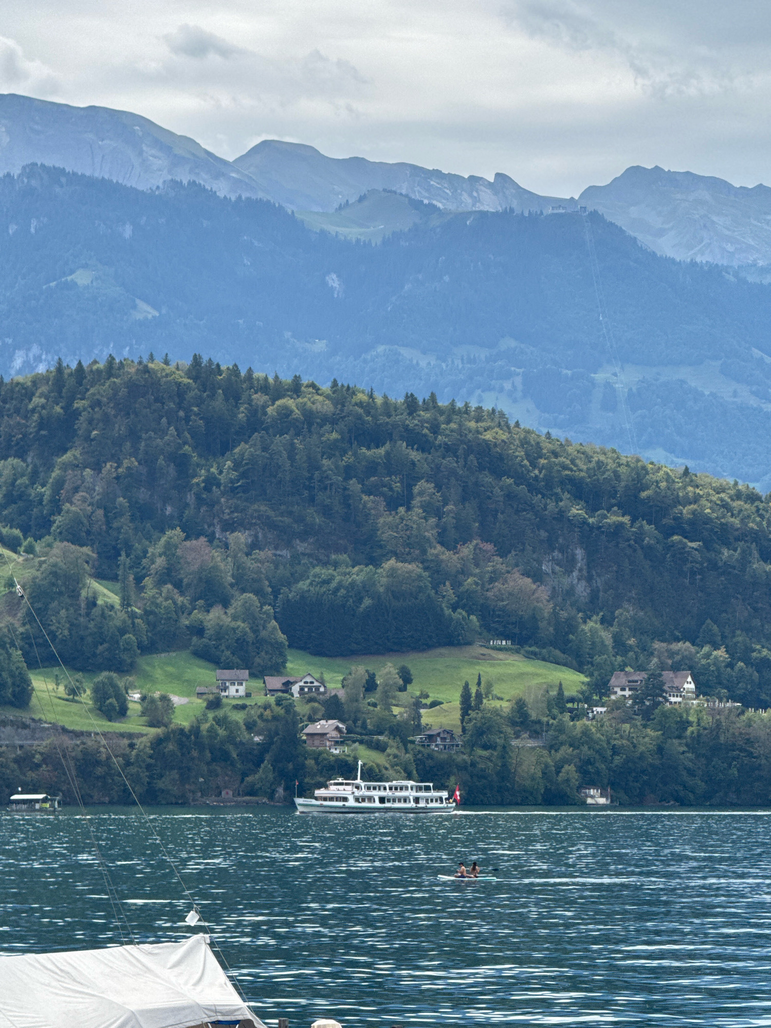 A wide shot captures a serene lake scene in Vitznau, Switzerland, with lush green mountains forming a dramatic backdrop. In the foreground, a large white boat is partially visible, its sail taut against the sky. The dark blue water of the lake is rippled, reflecting the overcast sky. Further out, a white passenger boat, flying a Swiss flag, glides across the water. Two figures are visible on a paddleboard in the middle distance, enjoying the lake. The middle ground is dominated by a densely forested mountainside dotted with a few houses and green clearings. The background features layers of majestic blue-hazed mountains under a cloudy sky. A prominent cable car track is visible ascending one of the distant peaks, suggesting a popular tourist destination.