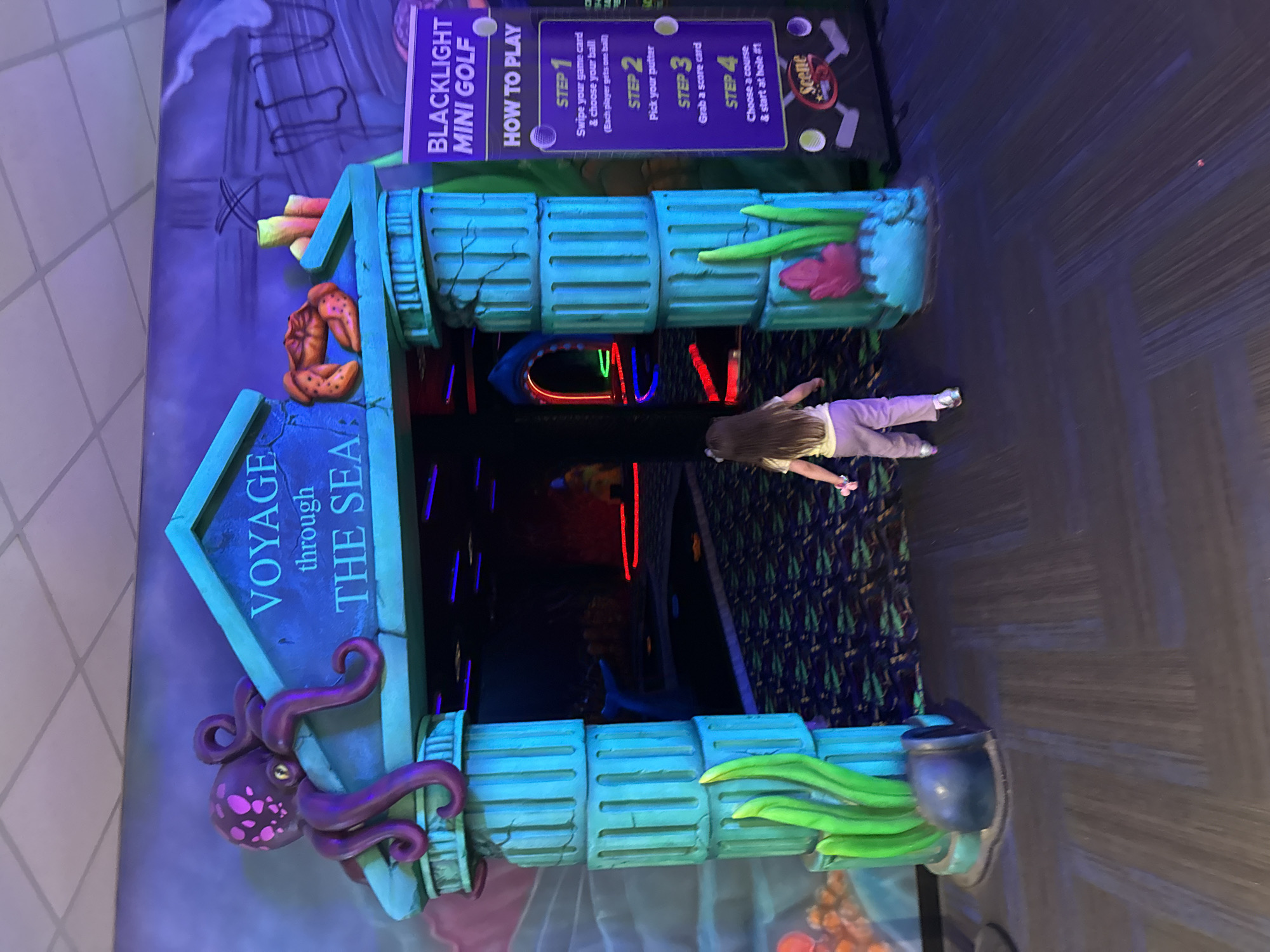 The image shows an entrance to a blacklight mini golf course called "Voyage through the Sea." The entrance is decorated with oceanic themes, including a large purple octopus on the left side, coral and other marine life on the top, and seaweed-like decorations on the right. A sign to the right details the steps for playing the game. A young girl is walking through the entrance. The setting appears to be indoors, with a dark, patterned floor and black walls, suggestive of a blacklight-lit environment. The mini golf course appears to be located in the city of Columbus, United States of America, based on information that would have been provided previously.