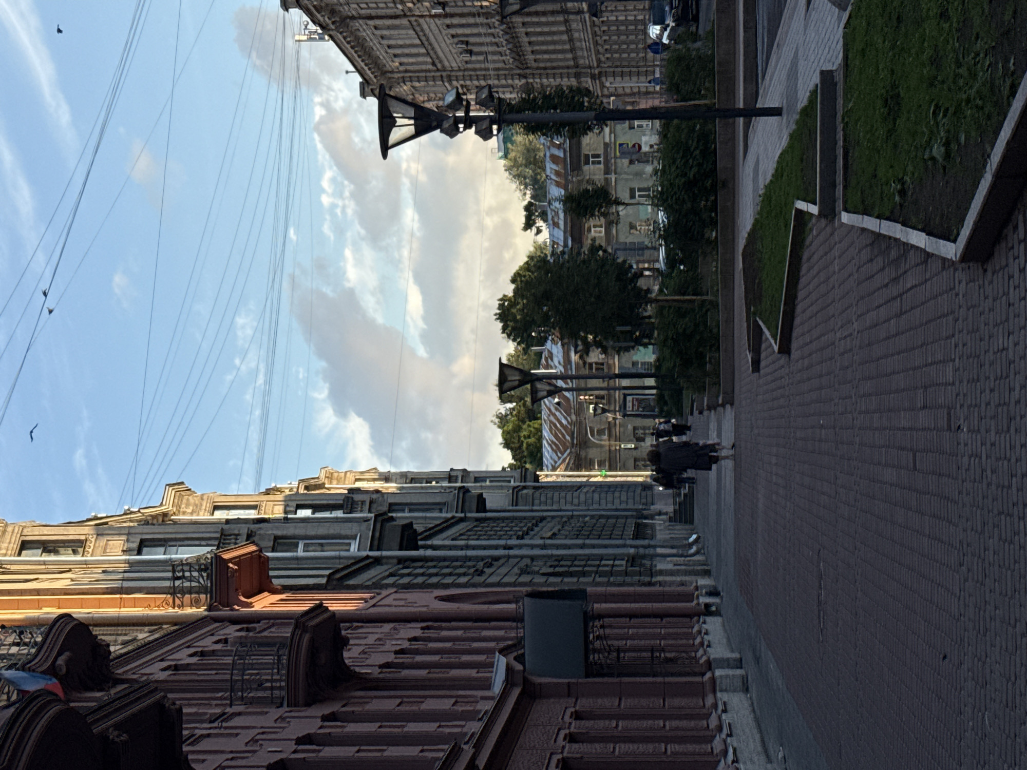 The image depicts a street scene in Saint Petersburg, Russia, likely in the early evening, given the lighting conditions. On the left side of the frame, a multi-story building with ornate balconies and a reddish-pink facade dominates the foreground. The building features arched windows and decorative architectural elements. To the right of this building, another structure with a grey facade is visible, extending further down the street.
The street itself is paved with red brickwork. Several people are walking along the sidewalk. In the middle ground, a person in a dark coat is walking away from the camera, accompanied by a smaller figure, possibly a child or a small dog. Further down the street, more figures can be seen walking.
The right side of the image shows a street with several lampposts. Beyond the street, more buildings are visible, including one with a corrugated metal roof. Trees and green spaces are interspersed along the street. The sky above is partly cloudy, with a few birds flying. Overhead, a complex network of electrical wires is visible. The overall atmosphere suggests a European city with historic architecture and a pedestrian-friendly streetscape.