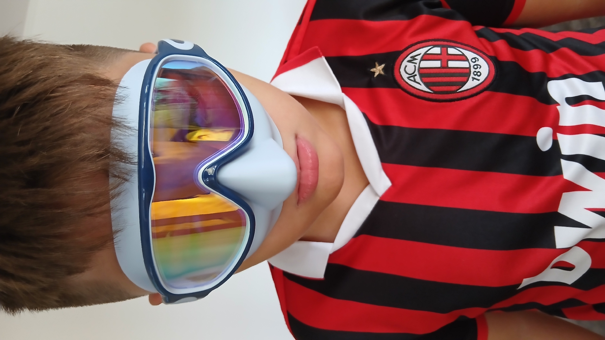 The image shows a close-up of a young person wearing snorkeling or swimming goggles and a red and black striped soccer jersey. The goggles are large, covering the eyes and nose, and have reflective lenses that show distorted colorful reflections. The jersey has a white collar and a crest on the left chest featuring "ACM" and "1899". There is also text on the jersey, partially visible, appearing to be "bwin". The background is plain and light-colored. The scene is indoors with soft lighting. This image is not from Pfarrkirchen, Germany.