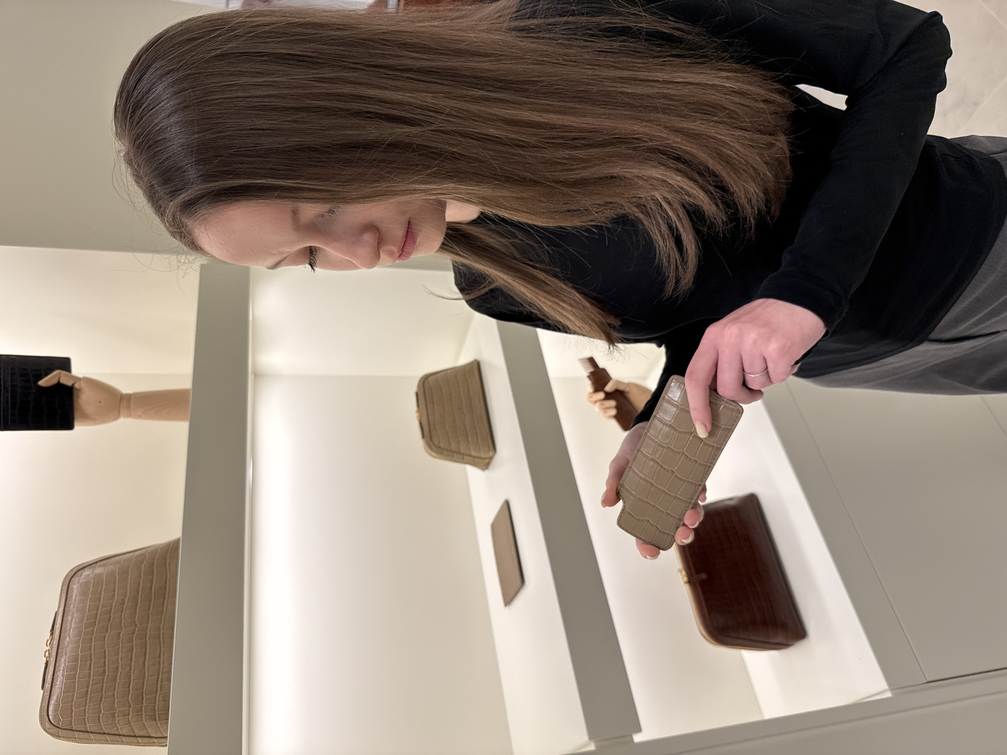This image, originating from Perm, Russia, depicts a female subject in a retail environment. The setting features a series of white display shelves with integrated under-shelf lighting, positioned against light-toned walls. On the uppermost visible shelf, a beige, crocodile-embossed zippered pouch is present, alongside a black, crocodile-embossed item held by a wooden articulated mannequin hand. The middle shelf displays a small, flat, beige, crocodile-embossed card holder and another beige, crocodile-embossed zippered pouch. On the lowest shelf, a dark brown, crocodile-embossed zippered pouch is visible. A female individual, characterized by long, straight brown hair and wearing a black long-sleeved top with grey trousers, occupies the right foreground. She holds a small, rectangular, beige-colored item with a crocodile-embossed texture in both hands, directing her gaze downwards to inspect the object. A thin silver ring is worn on her left ring finger.