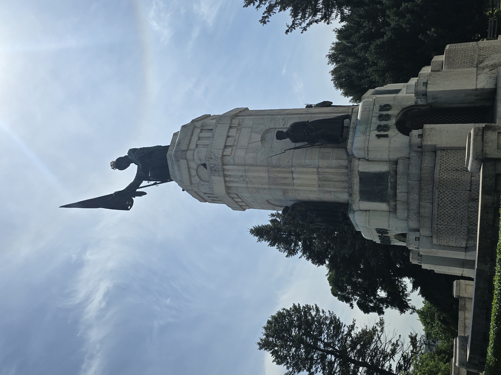The image shows a monument dedicated to Romanian heroes, likely in Bucharest, Romania, as the monument bears the date 1881, which is significant in Romanian history. The monument features several bronze figures, including one at the top with a flag, and several others around the base of the structure. The monument itself is constructed from stone or concrete and is a multi-tiered design. The background shows trees and a blue sky with some clouds, suggesting it's a sunny day.