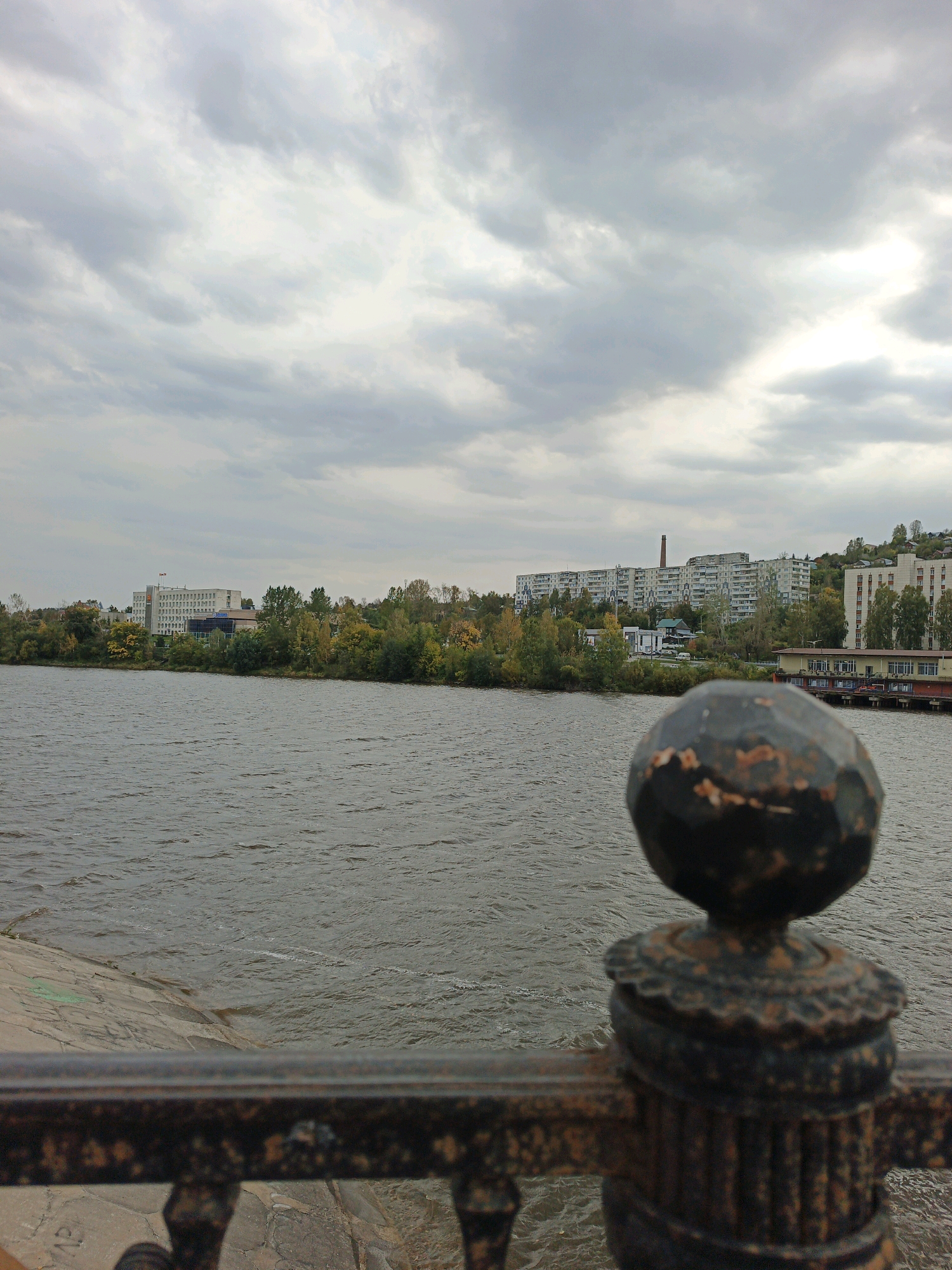 A wide, grey river stretches across the mid-ground of the image, with ripples on its surface. In the foreground, a dark, ornate metal railing is visible, out of focus, with a decorative post topped by a round, chipped finial. The background features a treeline with autumn foliage, behind which lie several multi-story apartment buildings. The sky is overcast with grey clouds. On the far left, a concrete embankment slopes down to the river. On the right side of the riverbank, a long, low building with a red roof is visible, possibly a pier or industrial structure. This scene appears to be located along the banks of the Ay River in Zlatoust, Russia. No people are visible in the image.