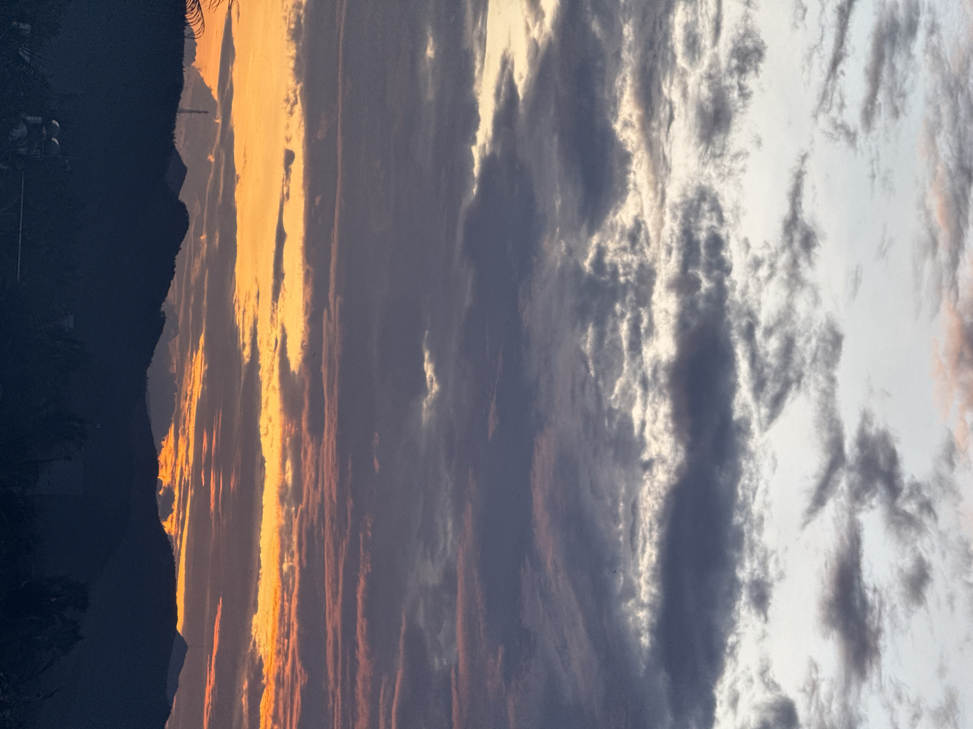 An atmospheric shot captures a dramatic sunset over a silhouetted mountain range. The sky is a canvas of shifting colors, with vibrant oranges and yellows bleeding into hues of pink, purple, and gray as the sun dips below the horizon. Dark, textured clouds drift across the sky, creating a sense of depth and drama. The mountains in the foreground are rendered as dark, imposing shapes, their forms softened by the fading light. A solitary radio tower stands prominent against the sunset, its structure starkly defined against the colorful sky. The lower portion of the image shows hints of foliage and structures, suggesting a populated area at the base of the mountains, but these details are largely obscured by shadow. The overall impression is one of natural beauty and serene, yet powerful, transition as day turns to night.