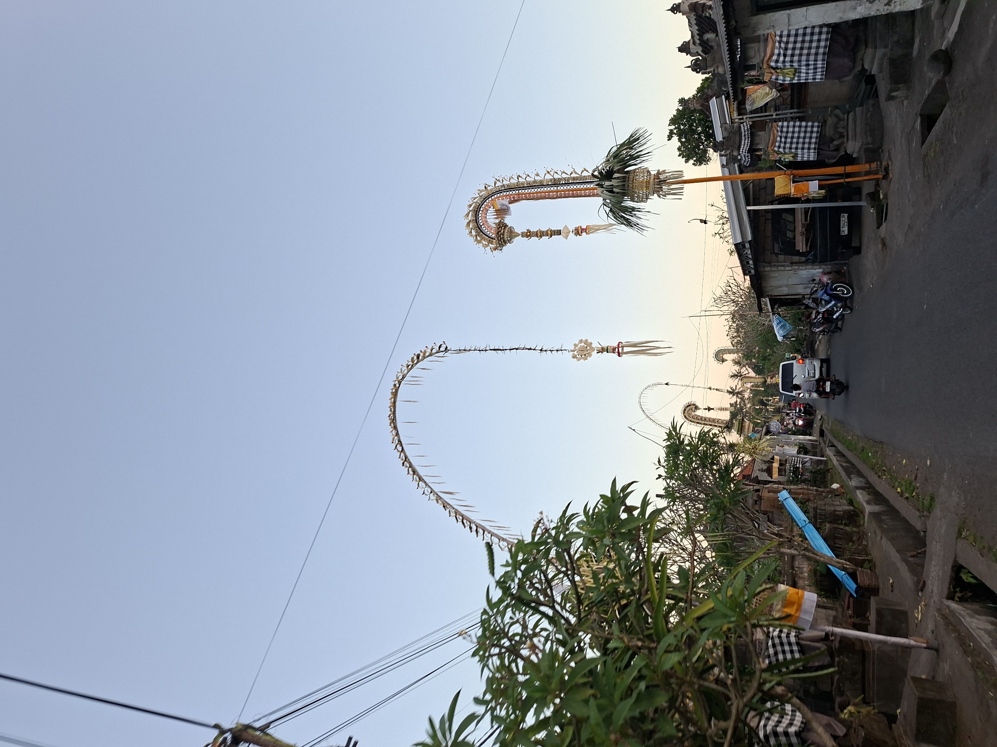 The image captures a street scene in Sukawati, Indonesia, likely during a festival or ceremony. Tall, arched decorations made of woven palm fronds and other natural materials adorn the sky, spanning across the road. These decorations are ornate and feature intricate patterns and hanging elements. On the left side of the street, lush green foliage from trees partially obscures the view of the buildings. Several traditional Balinese offerings or decorations are visible near the buildings, including checkered black and white cloths draped over stone structures.
The road itself is paved and appears to be a thoroughfare. A white van is seen driving away from the viewer, and several motorcycles are parked or also in motion. The buildings lining the street are a mix of simple structures, some with corrugated metal roofs, and more traditional stone buildings with ornate carvings. The sky is a clear, pale blue, indicating it is either early morning or late afternoon. The overall atmosphere suggests a blend of everyday life and cultural celebration.