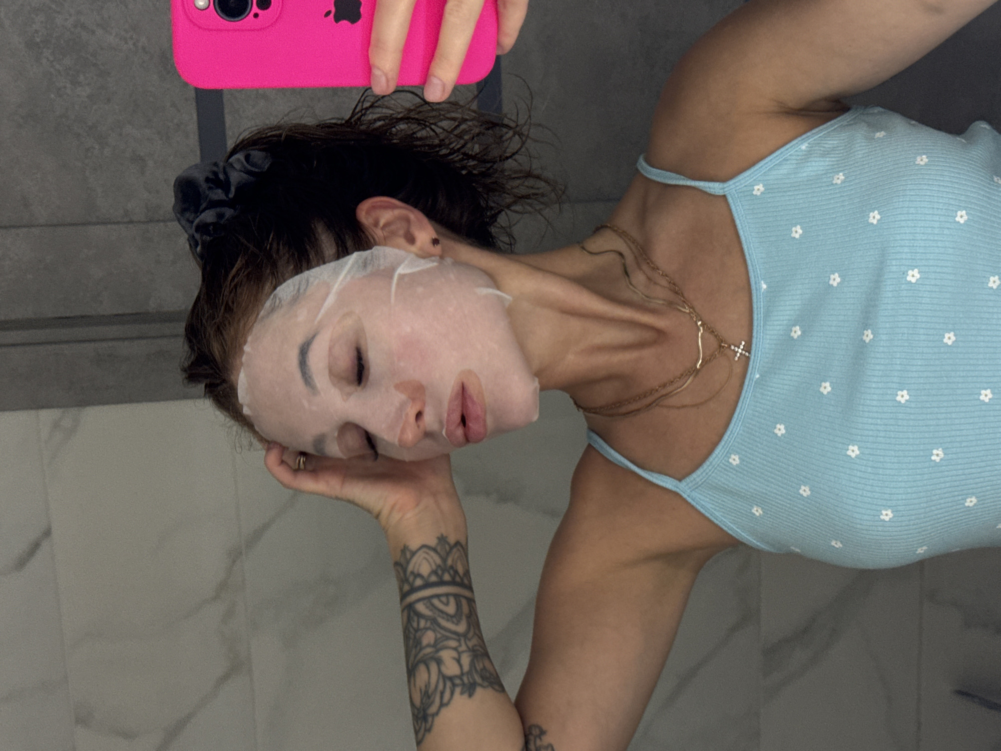 The image presents a close-up, rotated view of a person engaged in a self-care routine in Chaikovsky, Russia. The subject, likely a woman, has a white sheet mask covering her entire face. Her dark hair is pulled back with a black scrunchie and appears slightly damp. She is wearing a light blue ribbed tank top patterned with small white flowers and multiple gold-colored necklaces, one featuring a small cross pendant.
A prominent black tattoo with intricate, mandala-like patterns covers her left forearm and wrist. Her left hand is gently placed beneath her chin, supporting her head, while her right hand holds up a bright pink smartphone, identified as an iPhone by the visible Apple logo on its back, suggesting she is taking a selfie. Her eyes are closed, and her lips are slightly parted, conveying a sense of calm and relaxation during the skincare application.
The background consists of light gray or white tiled walls with a subtle marble-like pattern, indicating an indoor setting, likely a bathroom. The lighting is soft and even, highlighting the subject and her activity. The scene evokes a moment of personal pampering and tranquility.