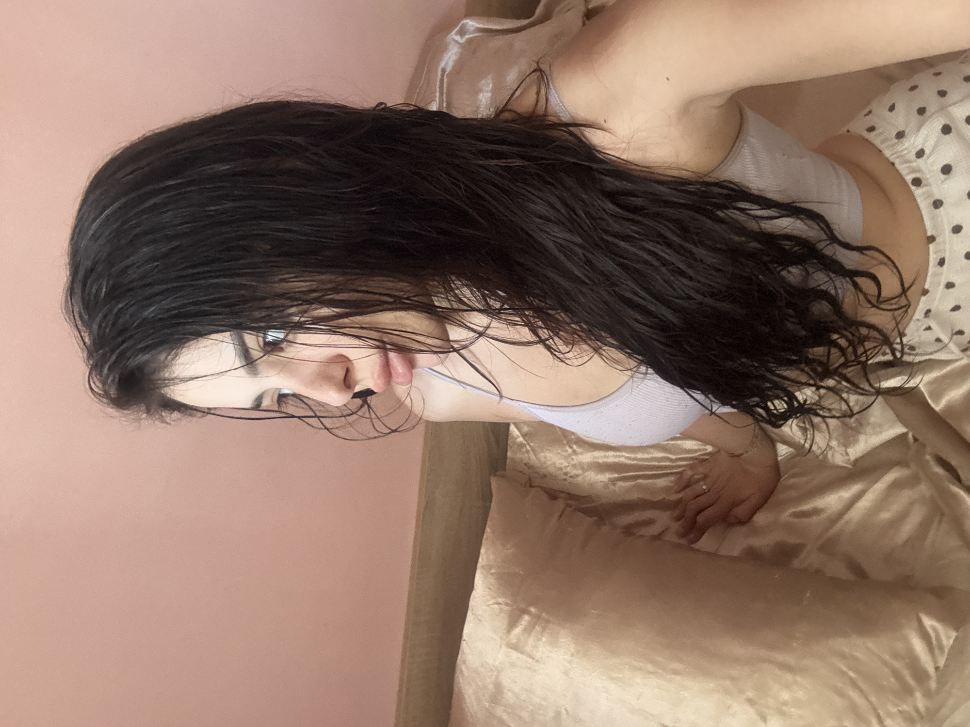 The image shows a woman lying on a golden-colored bed or couch. She has long, dark, wet hair and is wearing a light gray top. Part of her polka-dotted pants or shorts is visible. She is looking directly at the camera with a slightly downward gaze. The background is a peach-colored wall. There is no indication of Plovdiv, Bulgaria, or any city.